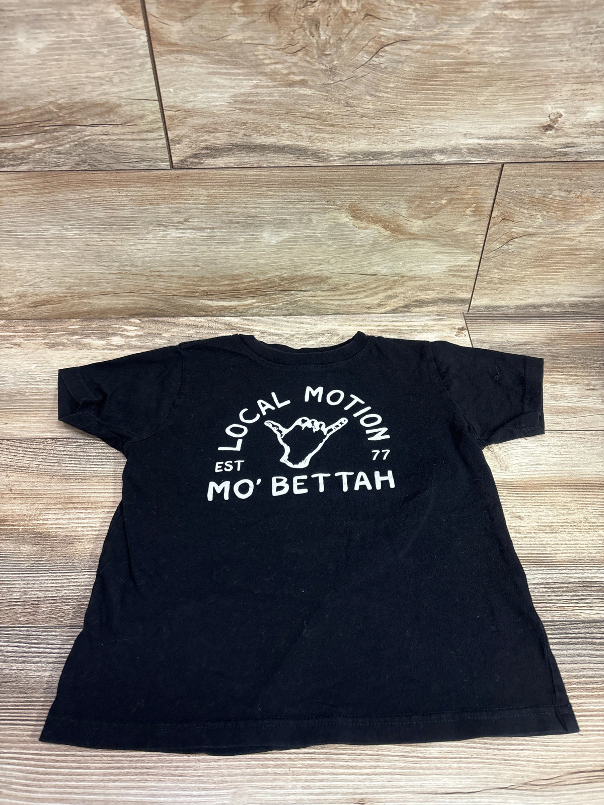 Front view of black Local Motion Hawaii shirt in size 5/6T showcasing white Mo’ Bettah graphic on a wood-look background