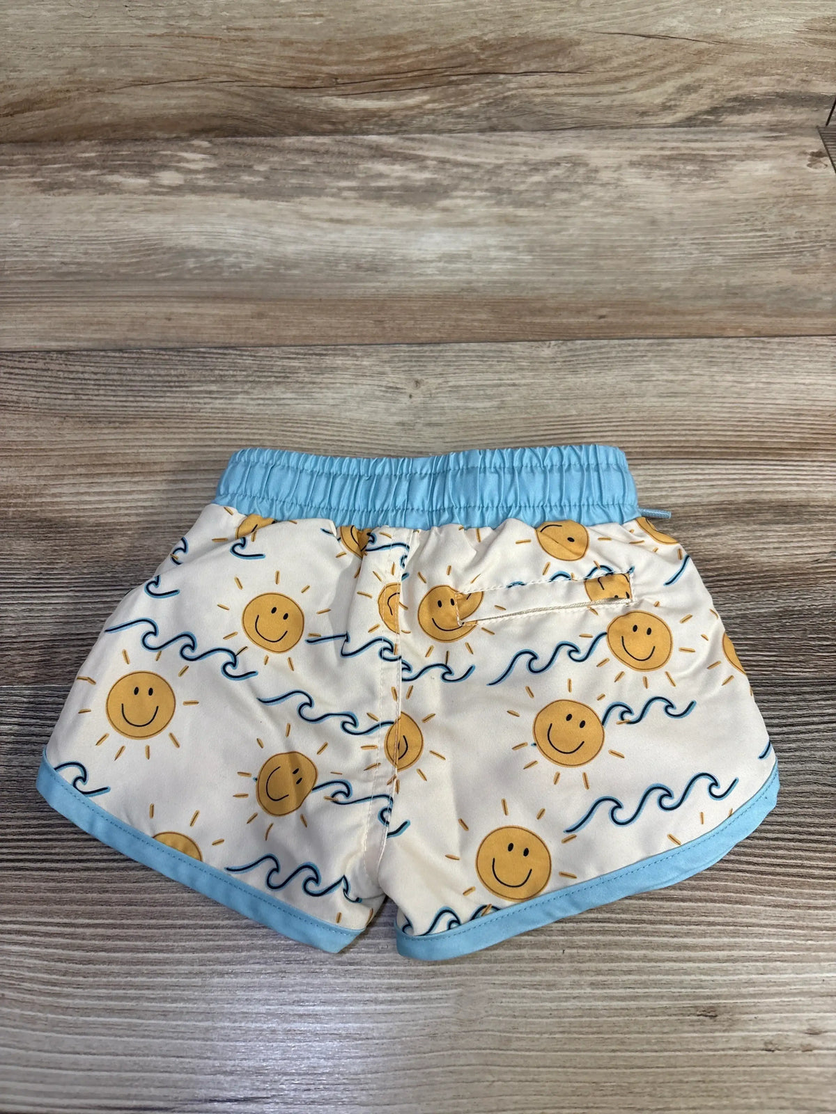 Flat lay of newborn swim trunks with cream base, yellow suns, blue wave print, elastic light-blue waistband on wood