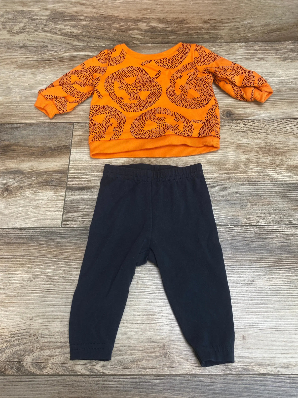Top-down view of Carter’s 2-piece jack-o-lantern set, orange long-sleeve shirt and black pants, size 3 months, on wood floor