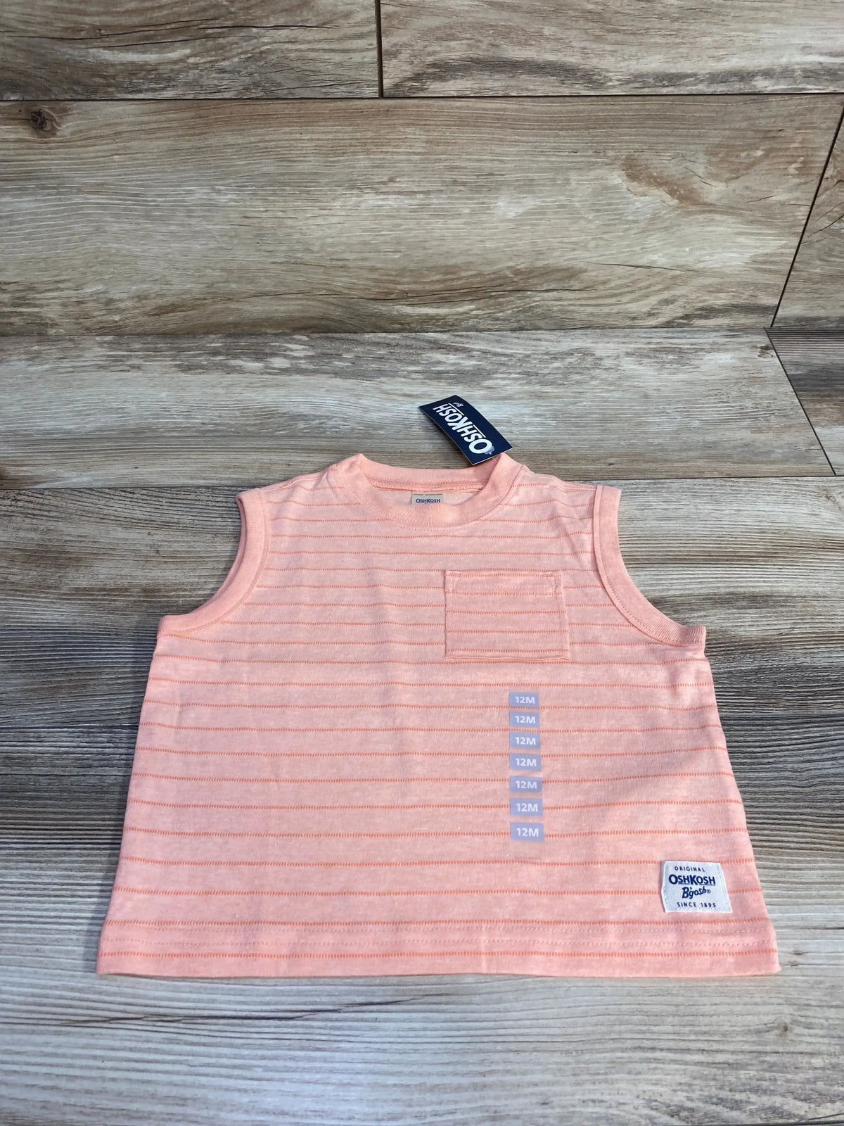 Top-down view of OshKosh pink striped baby tank top (12M) showing small chest pocket, label patch, and sleeveless cut on wood background