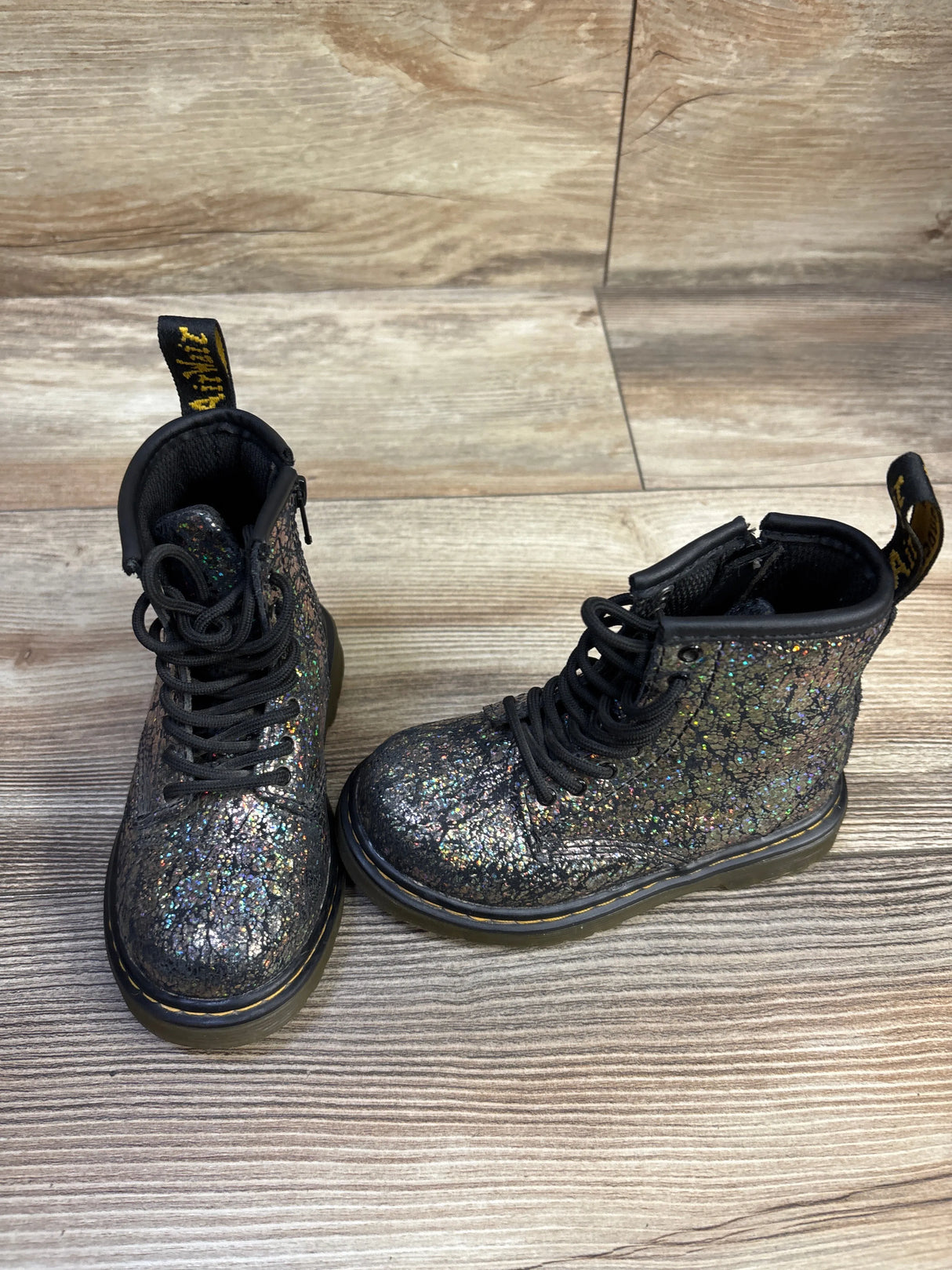 Three-quarter view of kids Dr Martens 1460 boots size 7C, black crinkle metallic glitter, lace-up with yellow welt on wood