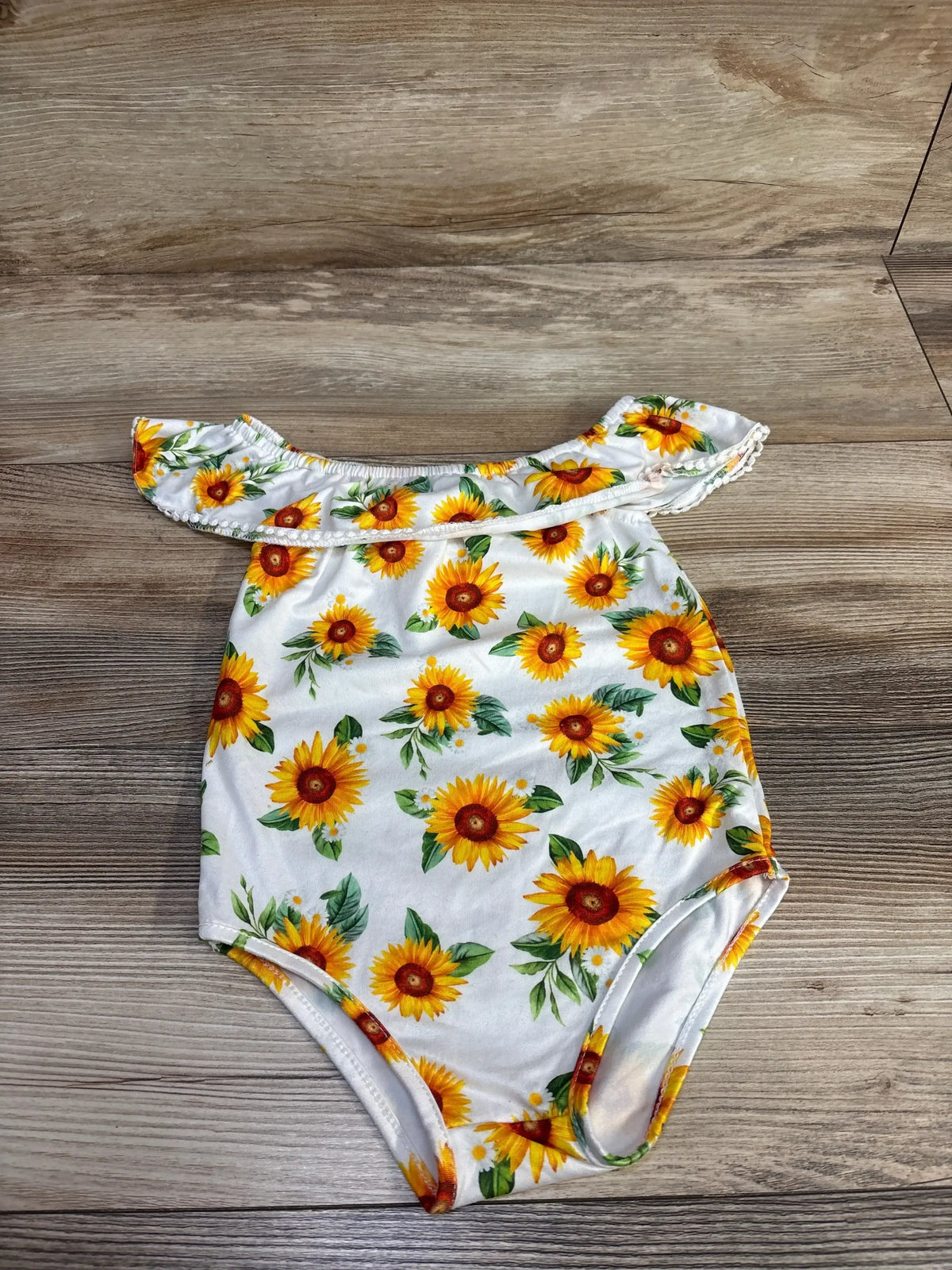 Flat lay of white baby off-shoulder romper showcasing ruffled neckline and yellow sunflower print on wood background