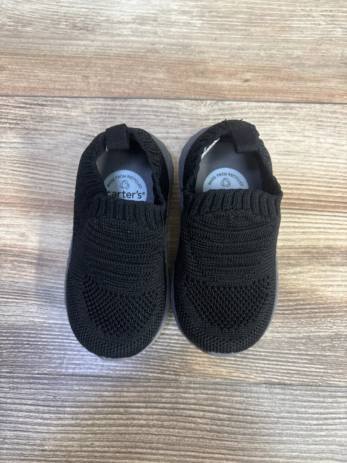 Top view of Carter’s Taylen pull-on athletic sneakers showcasing black knit uppers and elastic openings on a wood-look floor