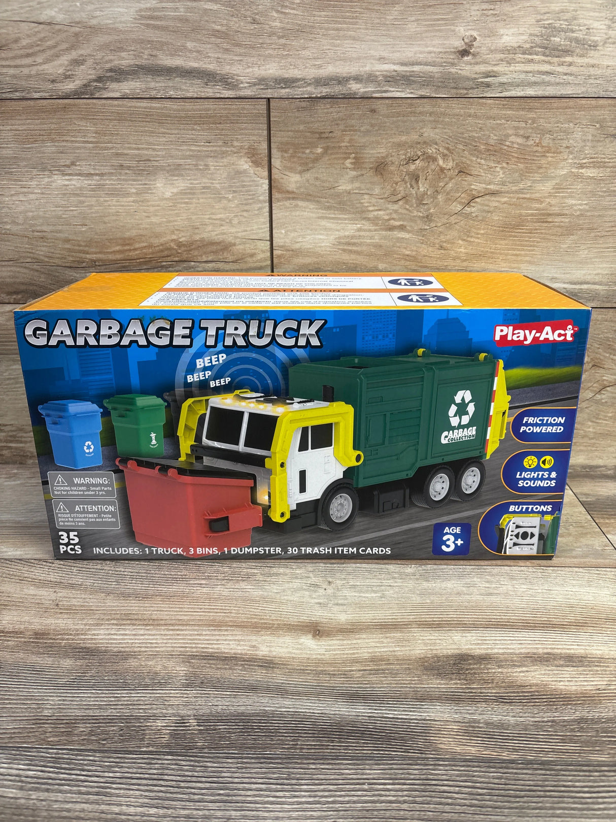 Three-quarter front view of Joyfy Play-Act 16-inch garbage truck toy in box showing green body, yellow cab, and red, blue, and green bins on wooden tabletop