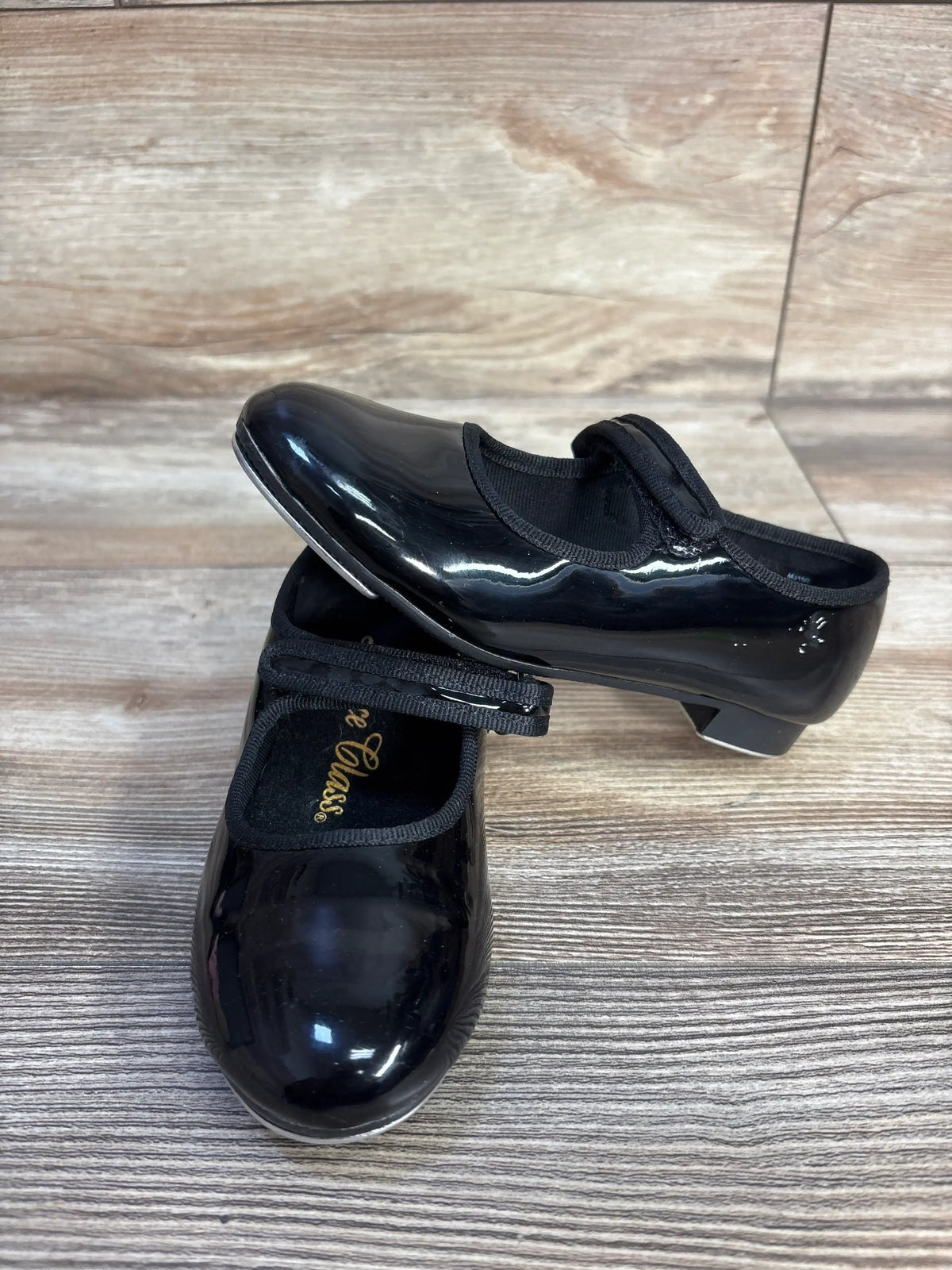 Pair of black patent leather children's tap dance shoes on a wood grain background.