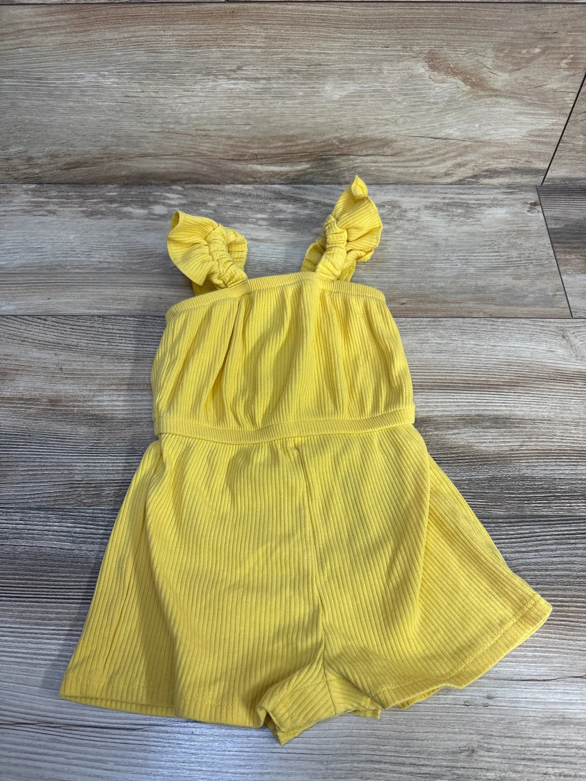 Front view of Old Navy ribbed tank shortie romper in yellow showcasing ruffled shoulder straps and shorts with a wood-look background
