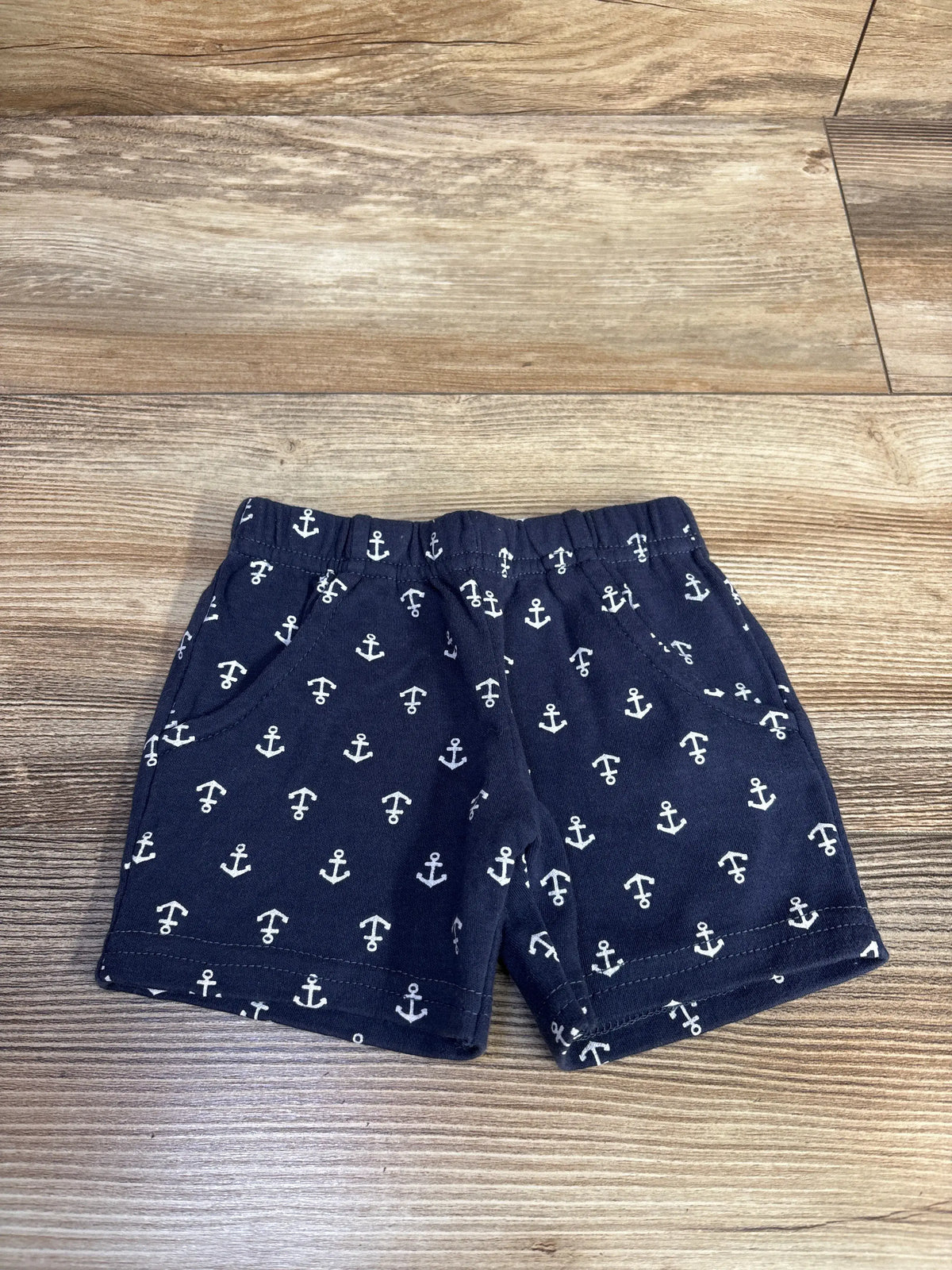 Front view of Hudson Baby anchor shorts showcasing navy cotton fabric, anchor print, elastic waistband, and side pockets on a wood-grain surface