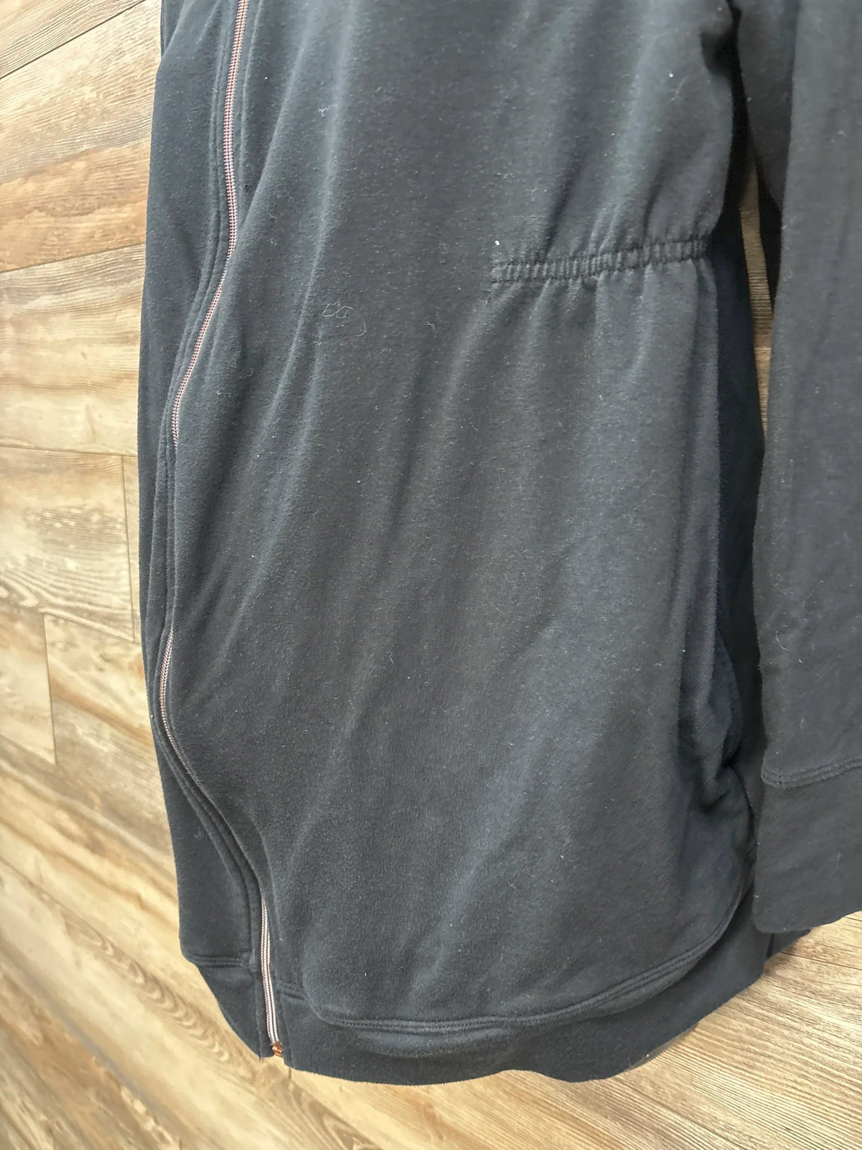 Side close-up of black Isabel maternity hooded zip-up jacket showcasing elasticated waist and full-length zipper against wood plank background