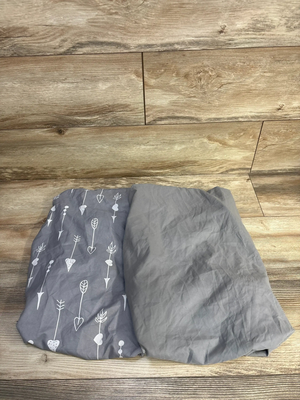 Top-down view of two fitted bassinet sheets, one solid grey and one grey with white arrow heart and leaf pattern on wood