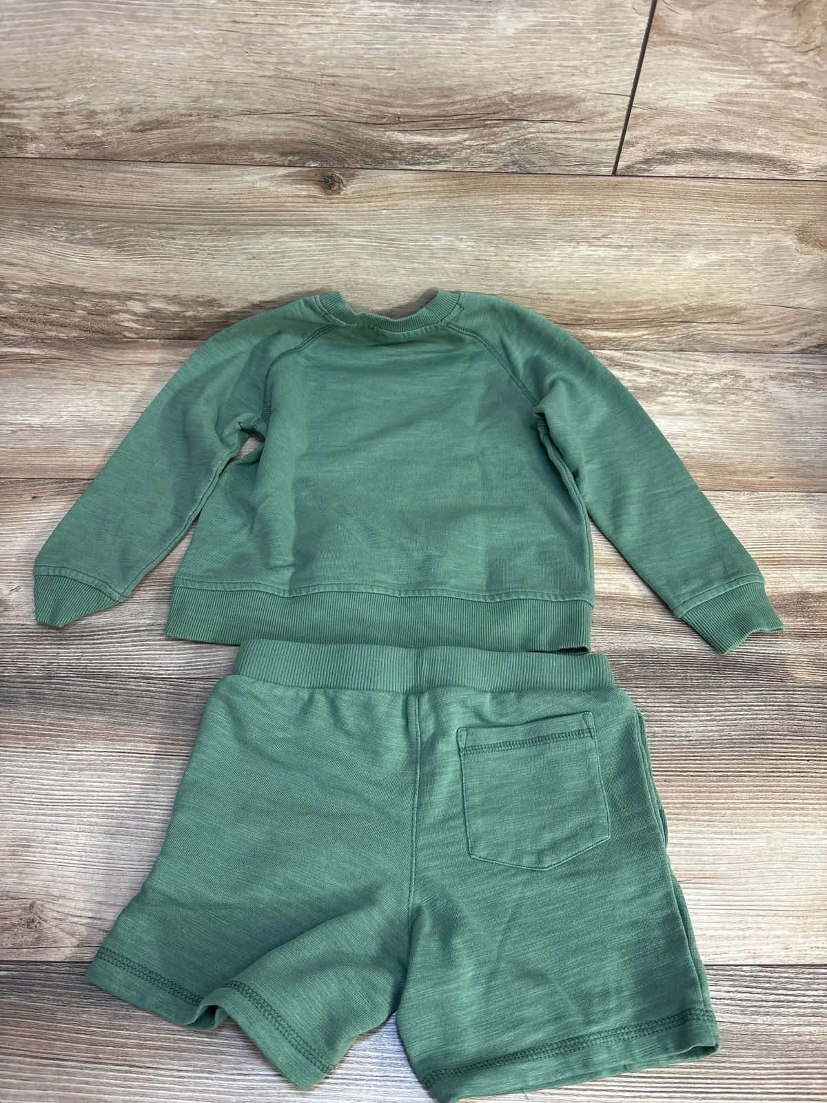 Flat lay of Janie and Jack green sweatshirt and shorts showing crewneck back and patch pocket on wood floor