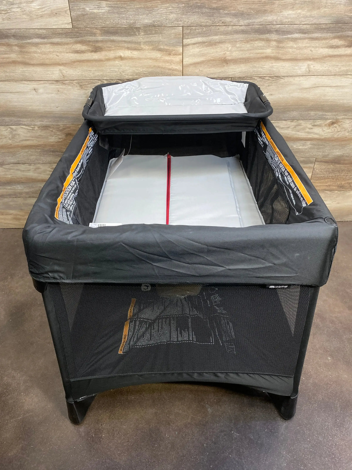 Front-facing view of 4moms Breeze Plus playard in black showing mesh sides, changing tray, and mattress against wood wall