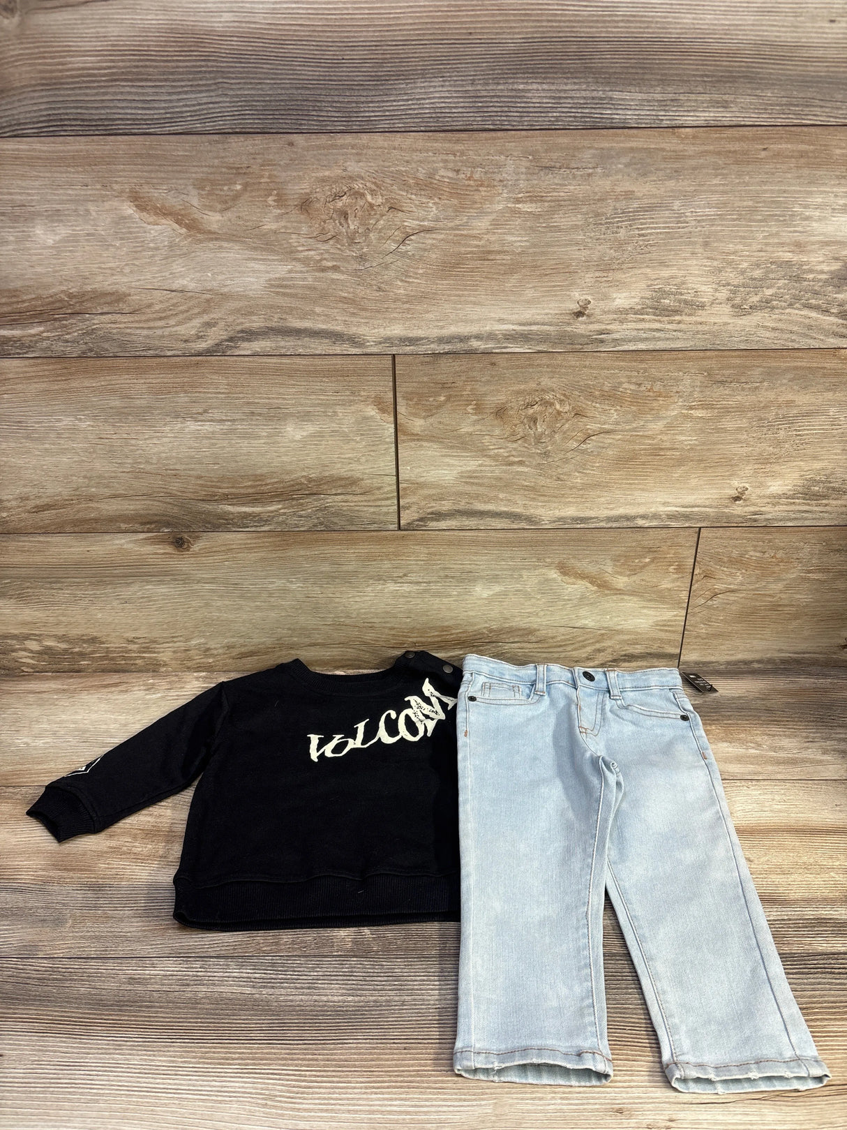 Flat front view of Volcom toddler 2-piece set, size 18 months, showcasing white logo on black long-sleeve shirt and light-wash jeans on wood background