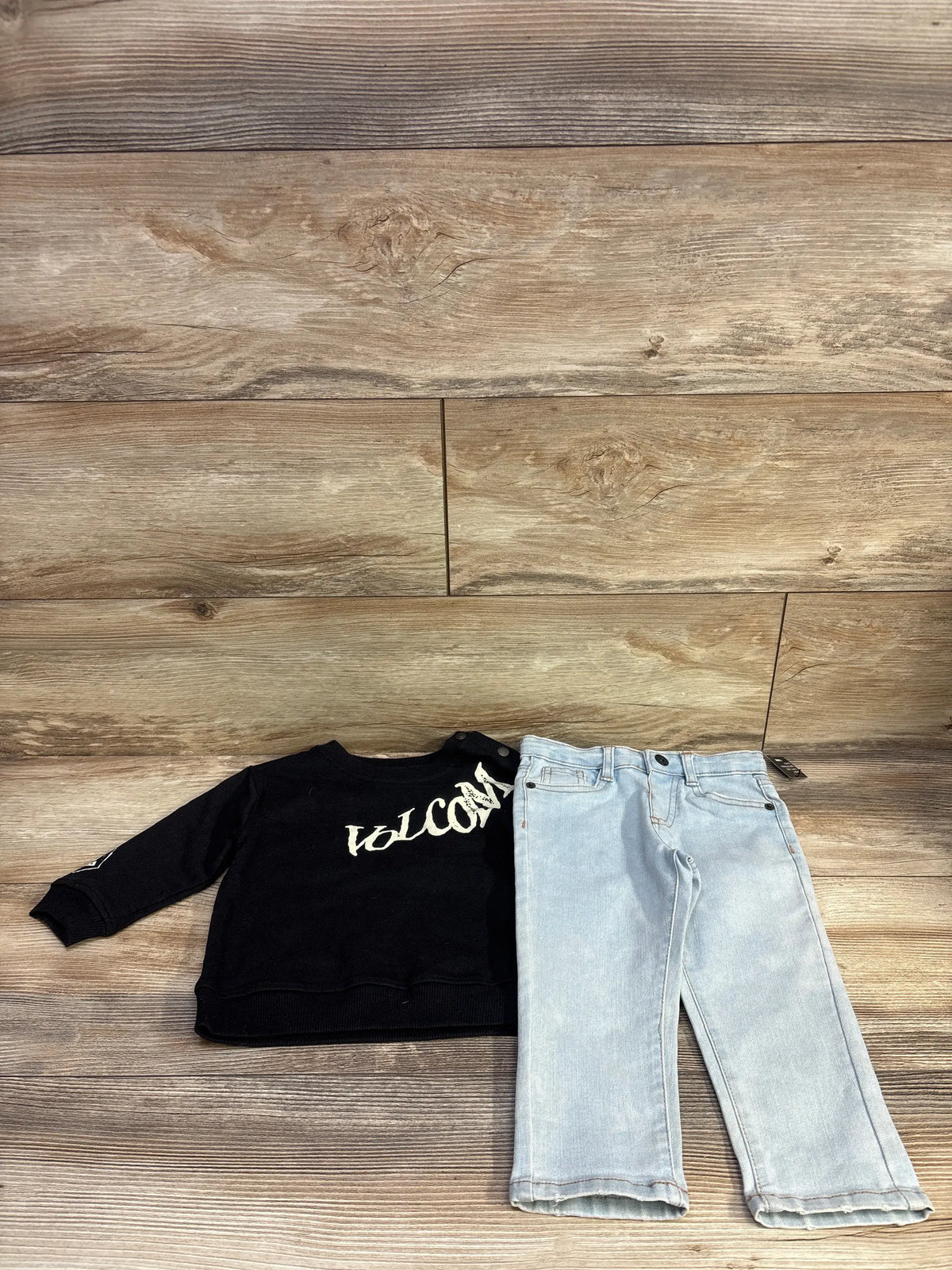 Flat front view of Volcom toddler 2-piece set, size 18 months, showcasing white logo on black long-sleeve shirt and light-wash jeans on wood background