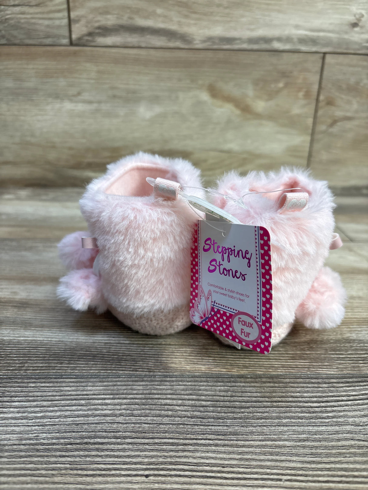Rear three-quarter view of pink faux-fur baby booties showcasing pom-pom accents and knit soles with wood plank background