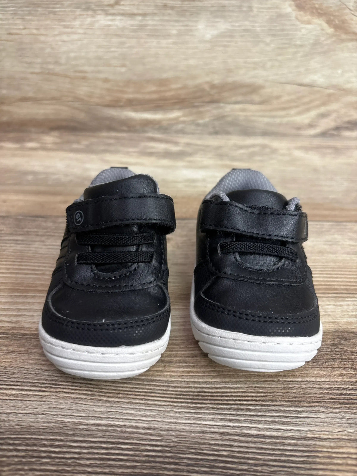 Front view of toddler Surprize by Stride Rite Killian black sneakers showing Velcro straps and white soles on wooden floor