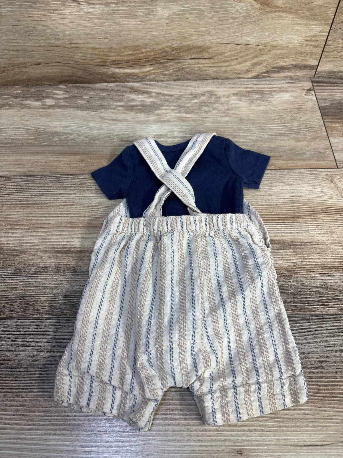 Top-down view of a newborn bodysuit and shortall set showcasing a navy bodysuit and blue-striped shortall on a wood floor