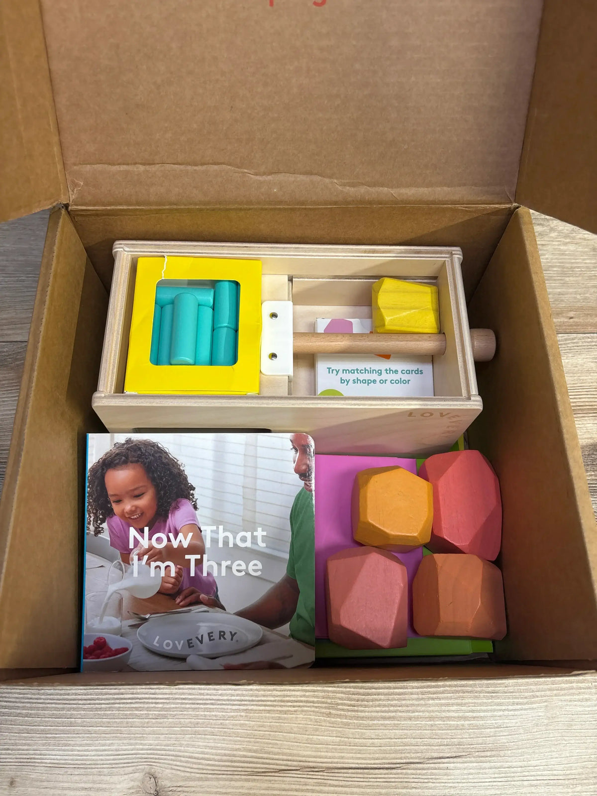 Top-down view of Lovevery Free Spirit Play Kit: wooden sorting box with hammer and teal pegs, four wooden gems, board book