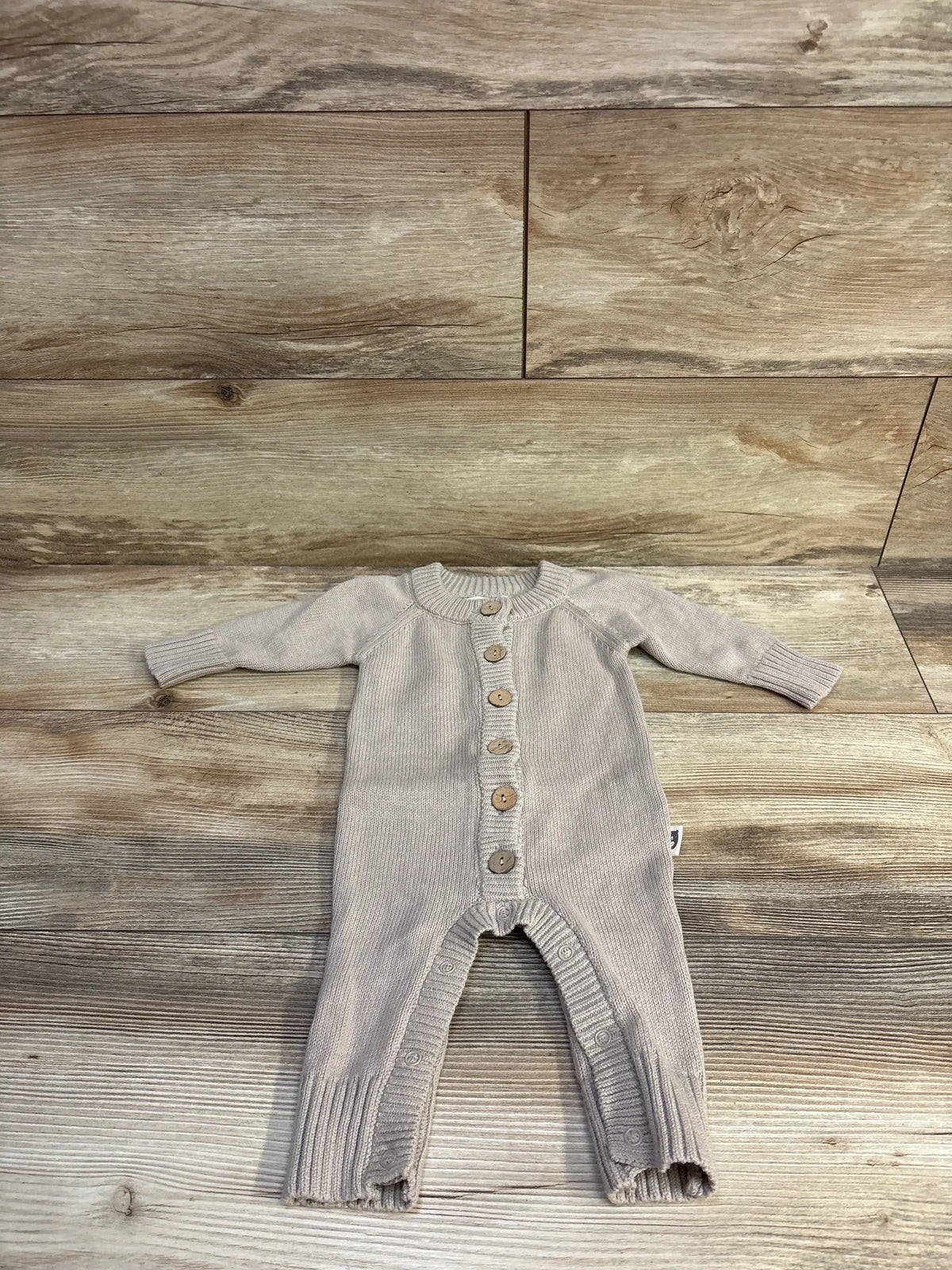 Flat-lay front of Rabbit + Bear organic knit coverall in light brown showing wooden buttons, ribbed cuffs, and leg snaps on a wood background