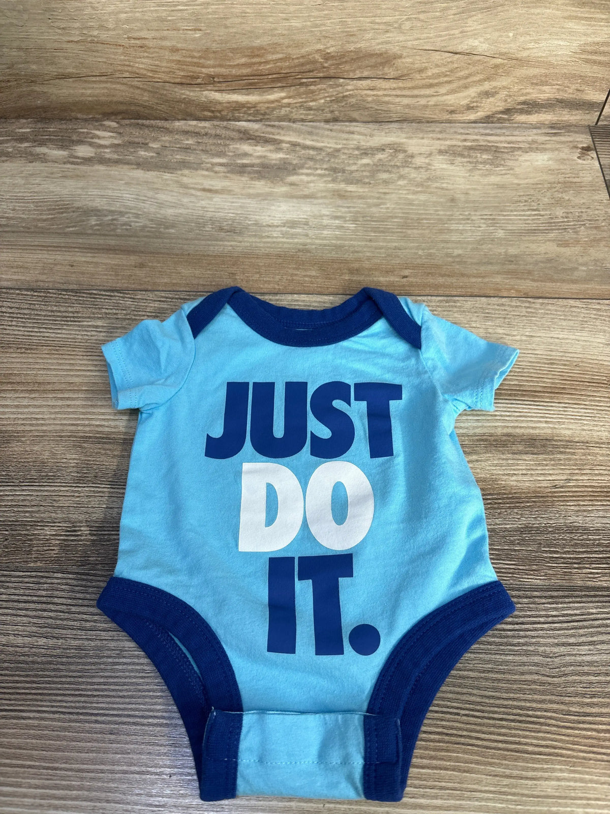 Flat lay of blue newborn Nike bodysuit showcasing large ‘JUST DO IT.’ front graphic, navy trim, and snap crotch with wood background