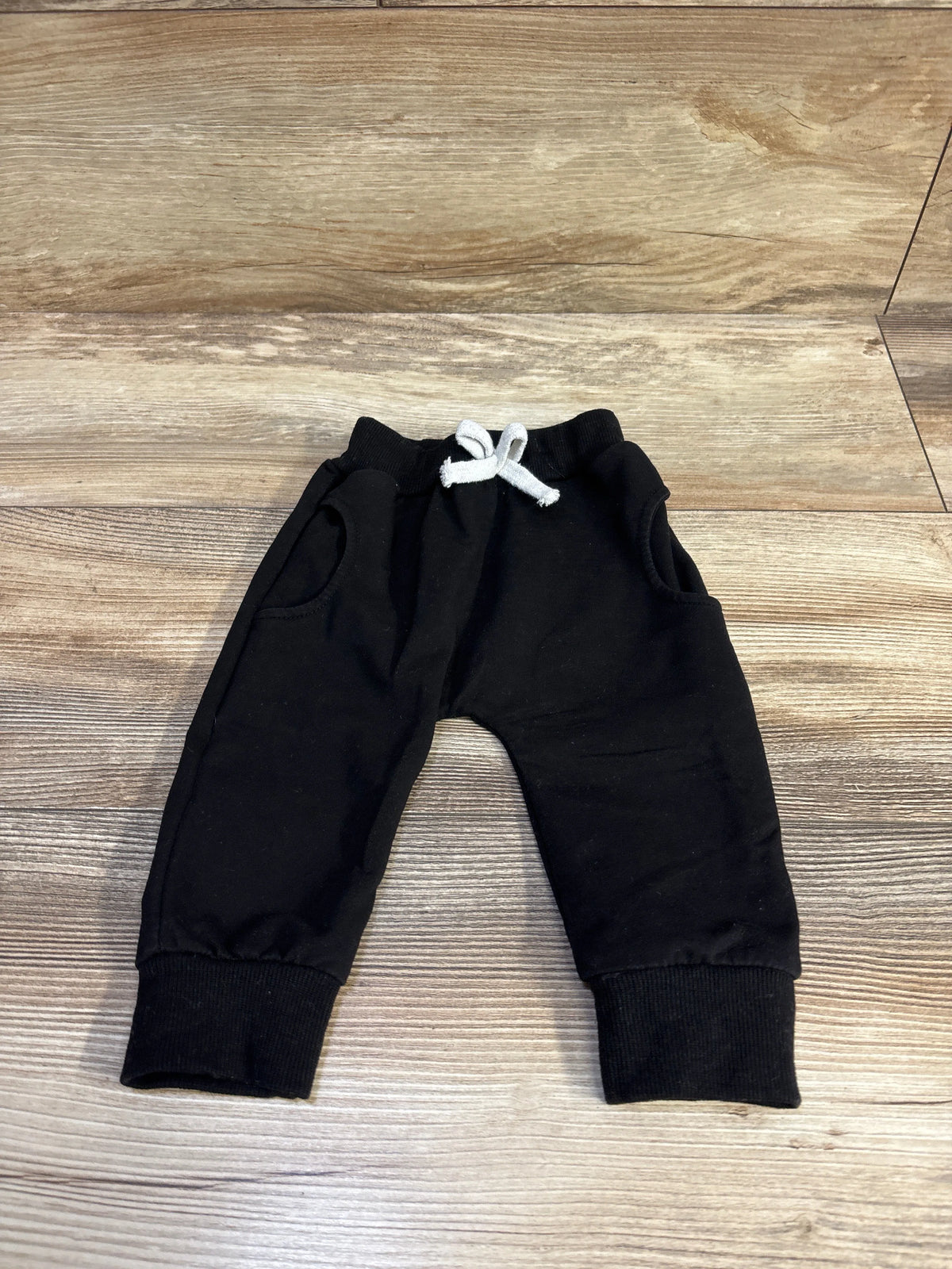 Front flat lay of black baby harem pants with white drawstring, ribbed ankle cuffs, and side pockets on wood background