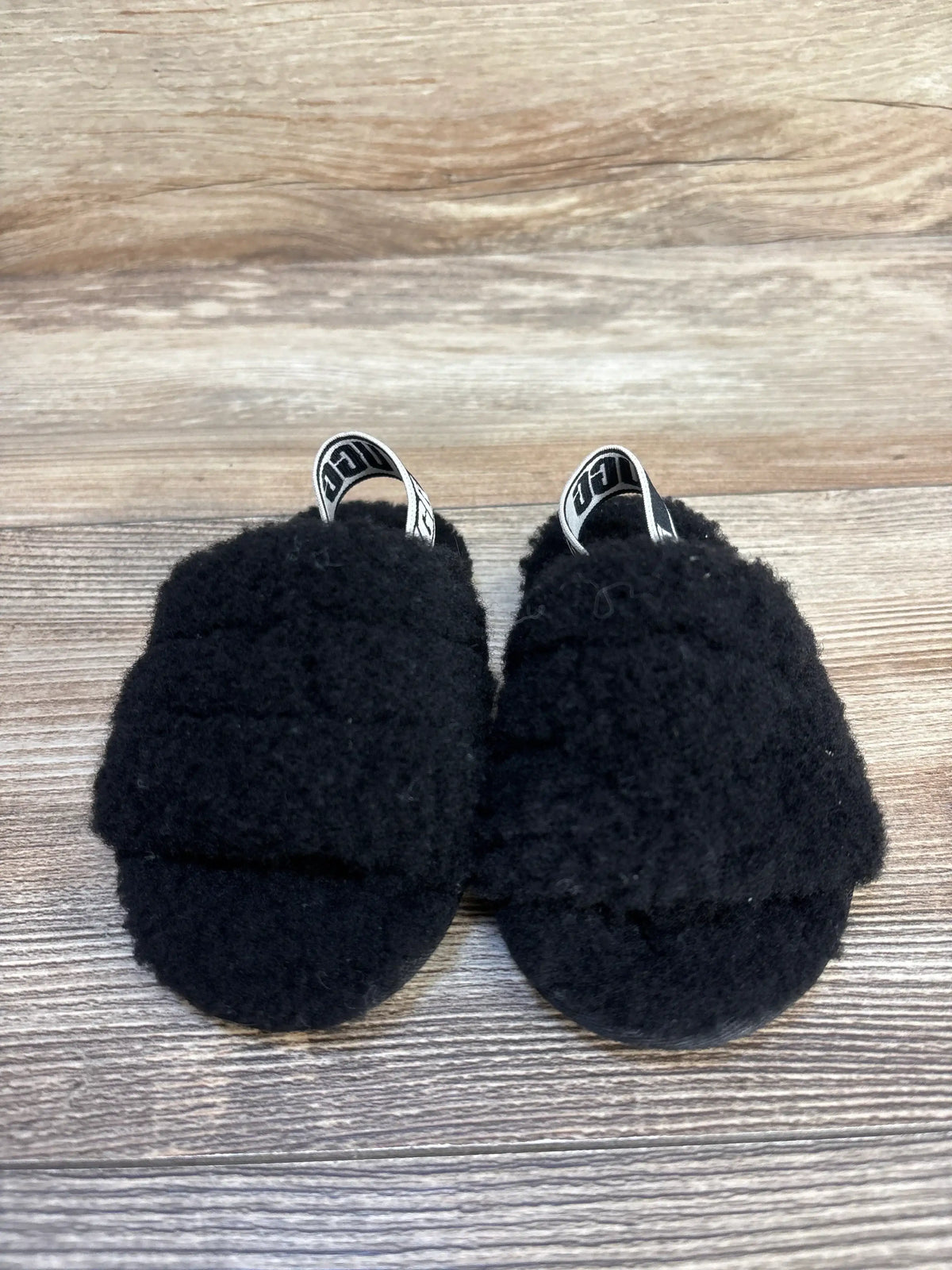 Top view of UGG Fluff Yeah baby slides in black showcasing fuzzy upper and elastic heel straps with UGG logo on wood background