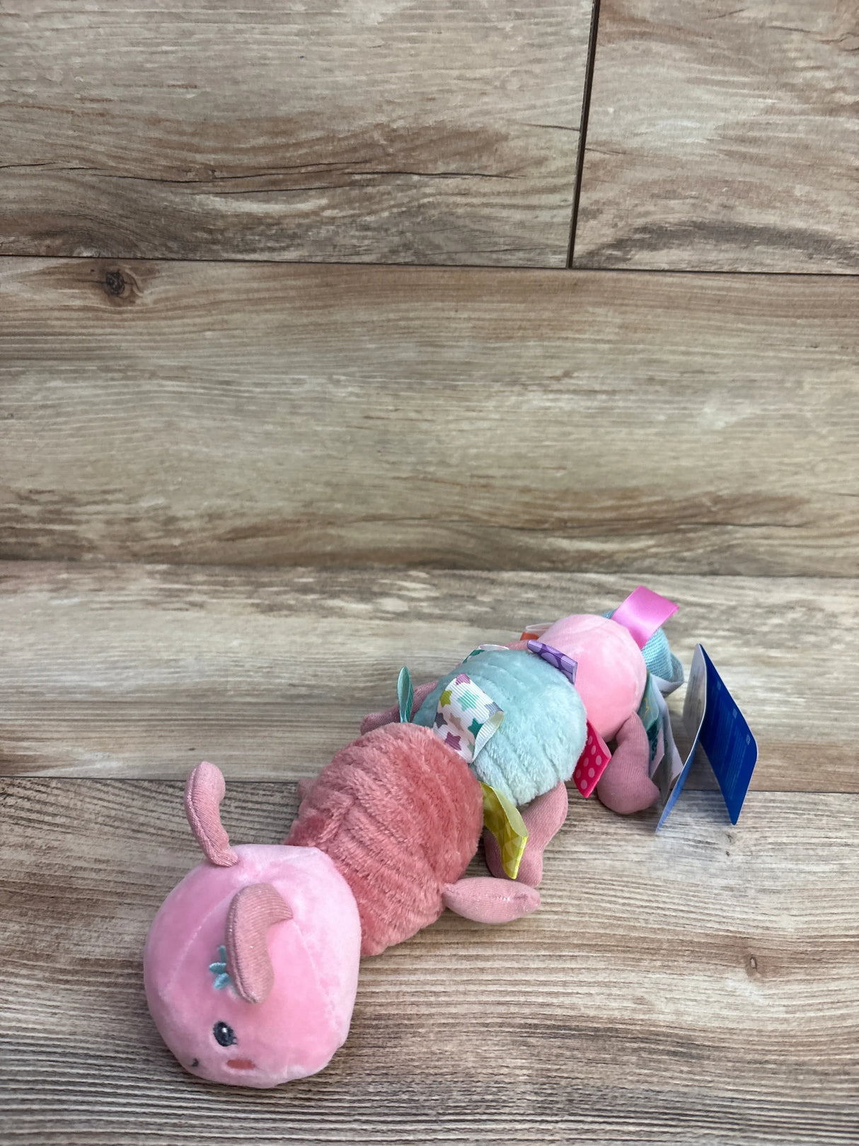 Plush pink caterpillar baby toy with colorful fabric tags on wood floor.