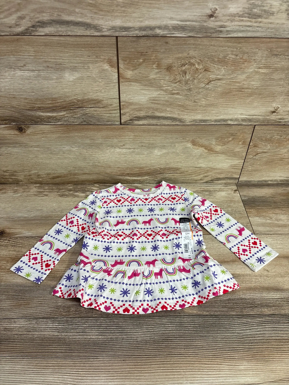 Front view of Okie Dokie long-sleeve peplum shirt with unicorn and rainbow fair isle print on wood background, size 12M tag visible