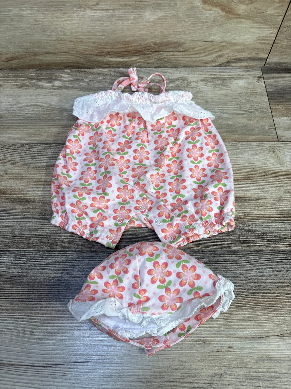 Top view of Charlotte & Star 2-piece tank romper and hat set showcasing pink floral print and white ruffled trim on a wood floor