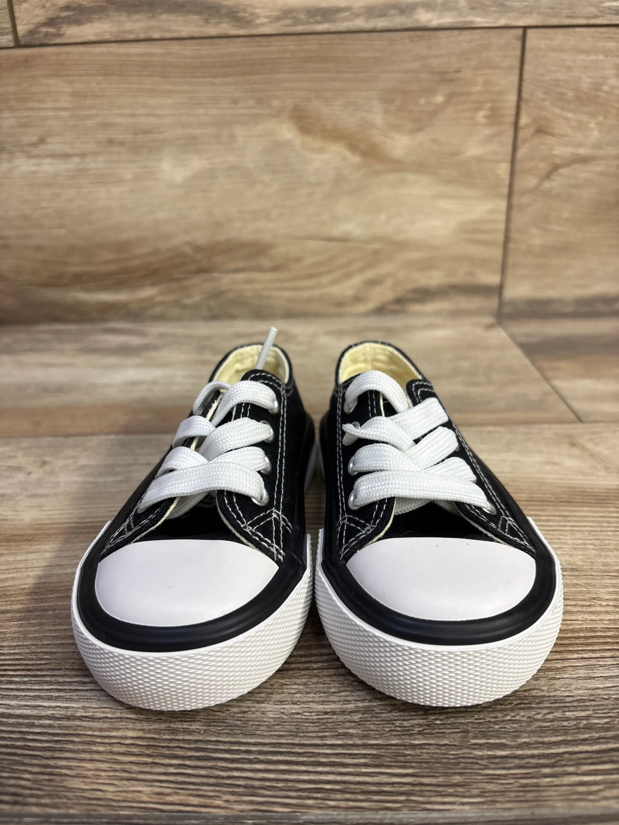 Front view of black toddler low-top sneakers size 5C showcasing white rubber toe caps, thick white soles, and white laces on wood floor