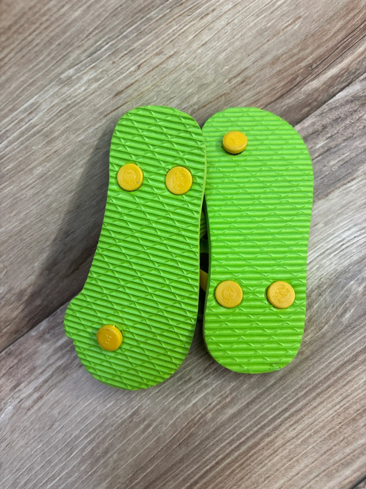Bottom view of lime green kids flip-flops showing textured soles and yellow strap plugs on wood floor, size 6-7