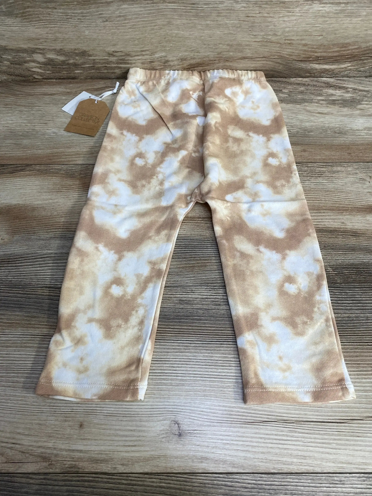 Flat lay of toddler tie-dye pants (size 2T) showing tan and white cloud pattern, elastic waistband and straight legs on wood floor