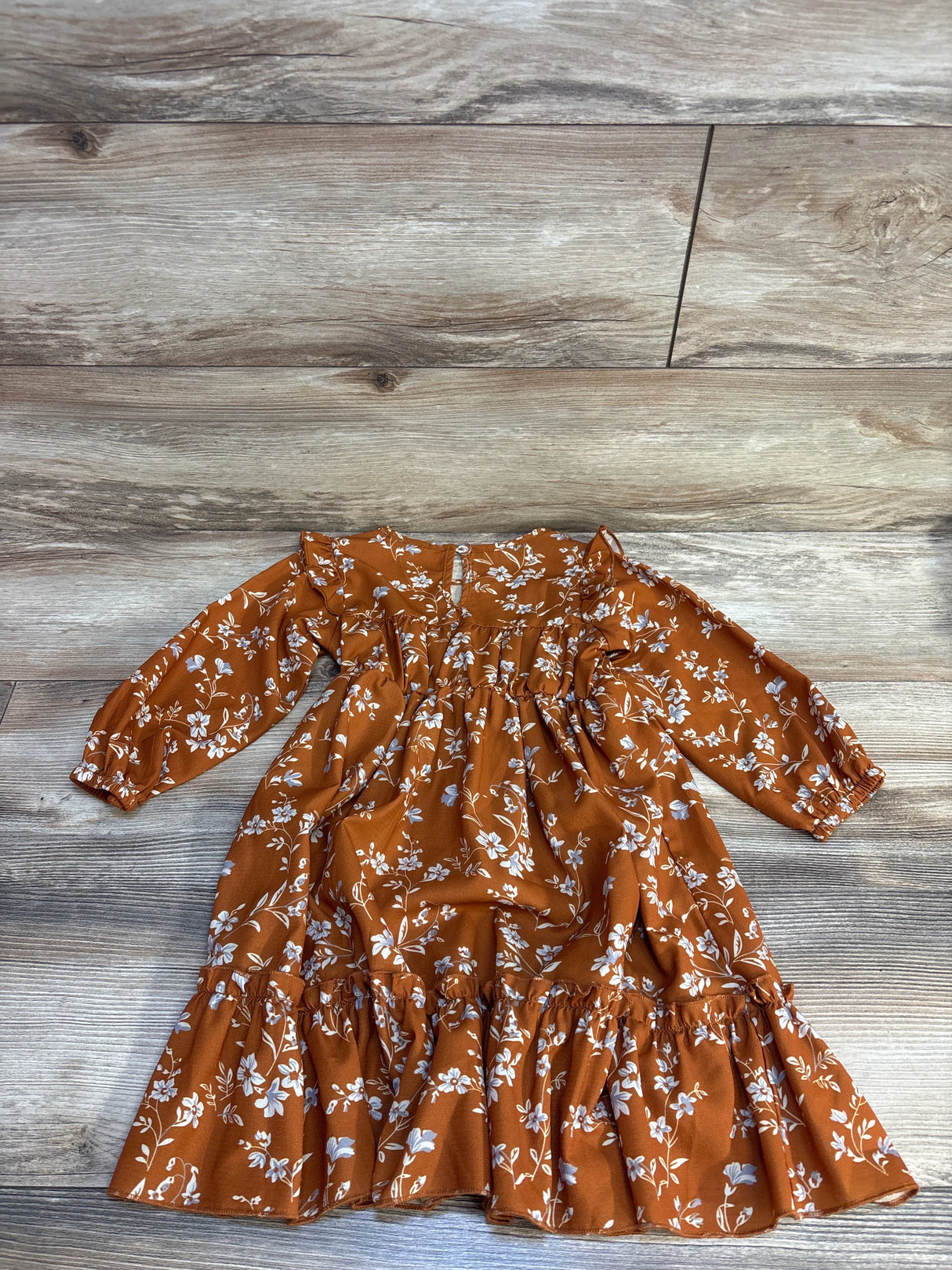 Flat lay of girls' long-sleeve ruffle dress, burnt orange floral, showing tiered hem and elastic cuffs on wood floor