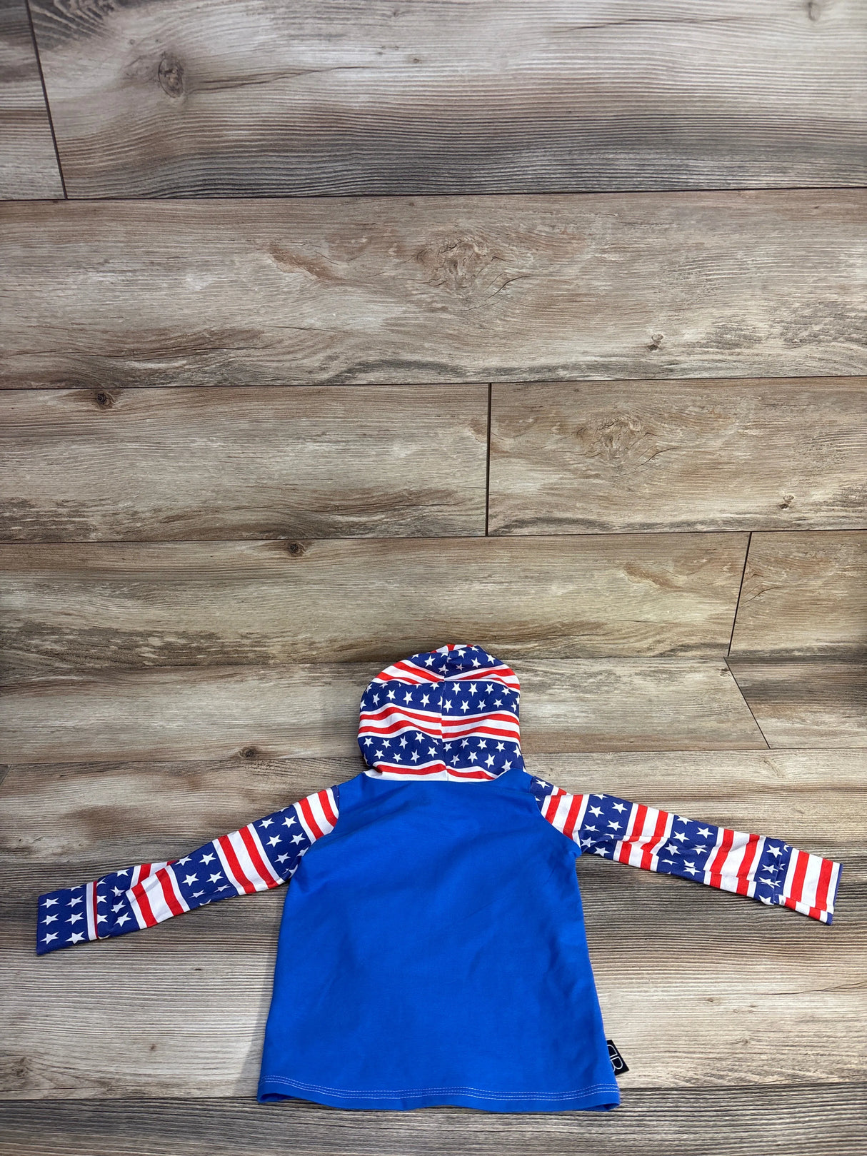 Back view of Baby Bums hooded rash guard with solid blue body and American-flag hood and sleeves, laid flat on wood background