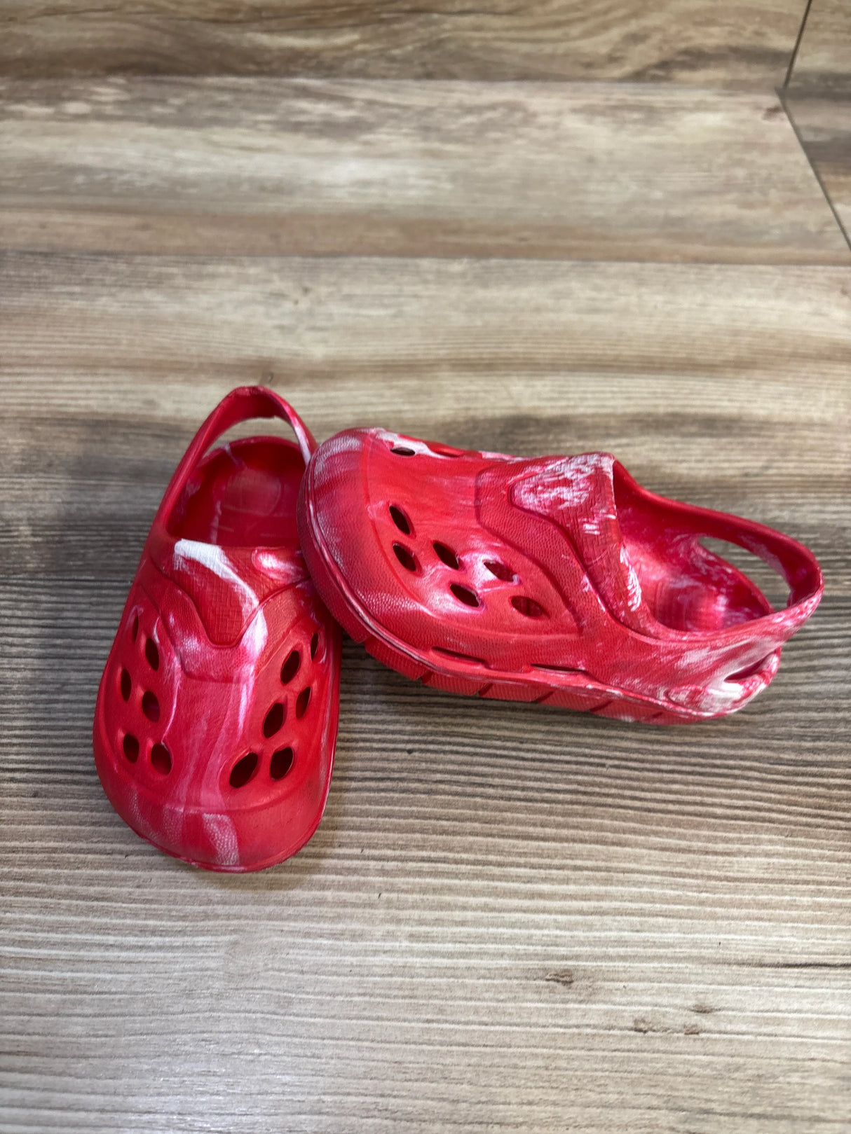 Three-quarter view of red tie-dye toddler clogs with white marbling, heel strap and ventilation holes on wood floor