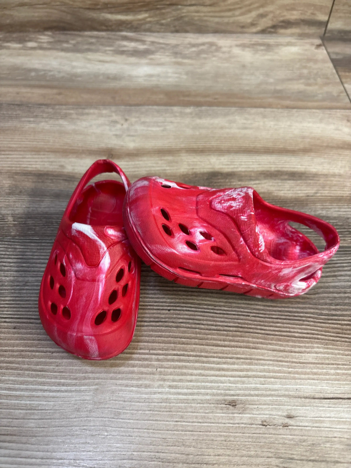 Three-quarter view of red tie-dye toddler clogs with white marbling, heel strap and ventilation holes on wood floor