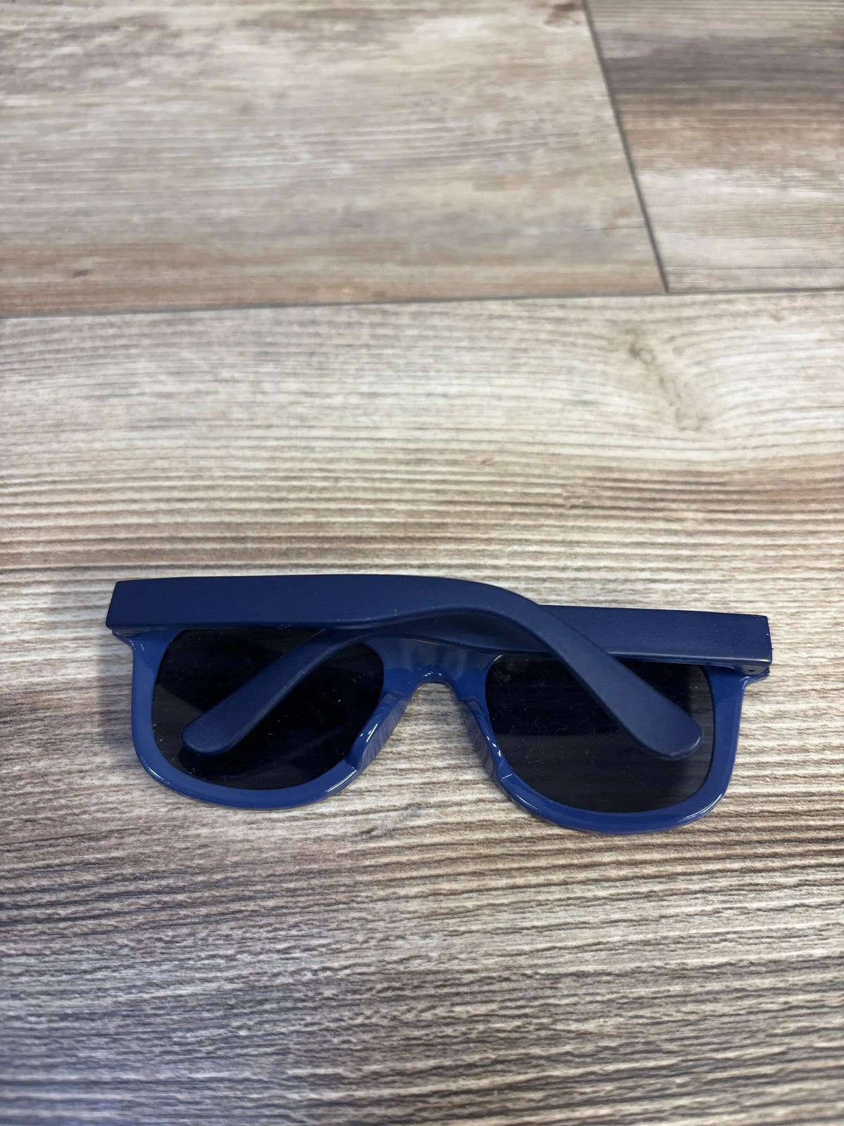 Top view of Just One You kids sunglasses in blue showing folded temples and dark lenses on a light wood surface