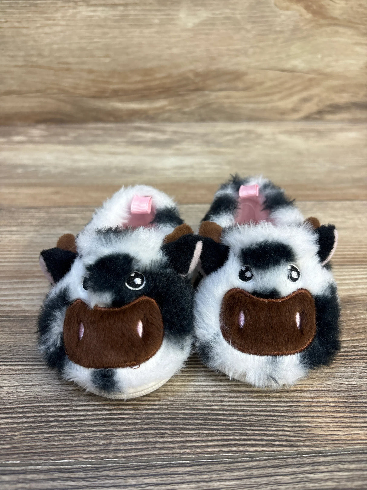 Plush baby slippers with black and white cow face design and pink bows on wooden floor
