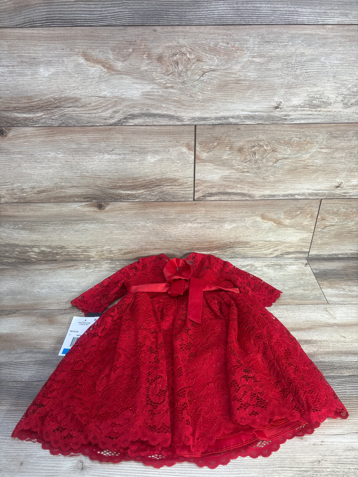 Flat lay of girls red glitter lace party dress showcasing long sleeves, bow at waist, and scalloped hem on light wood floor