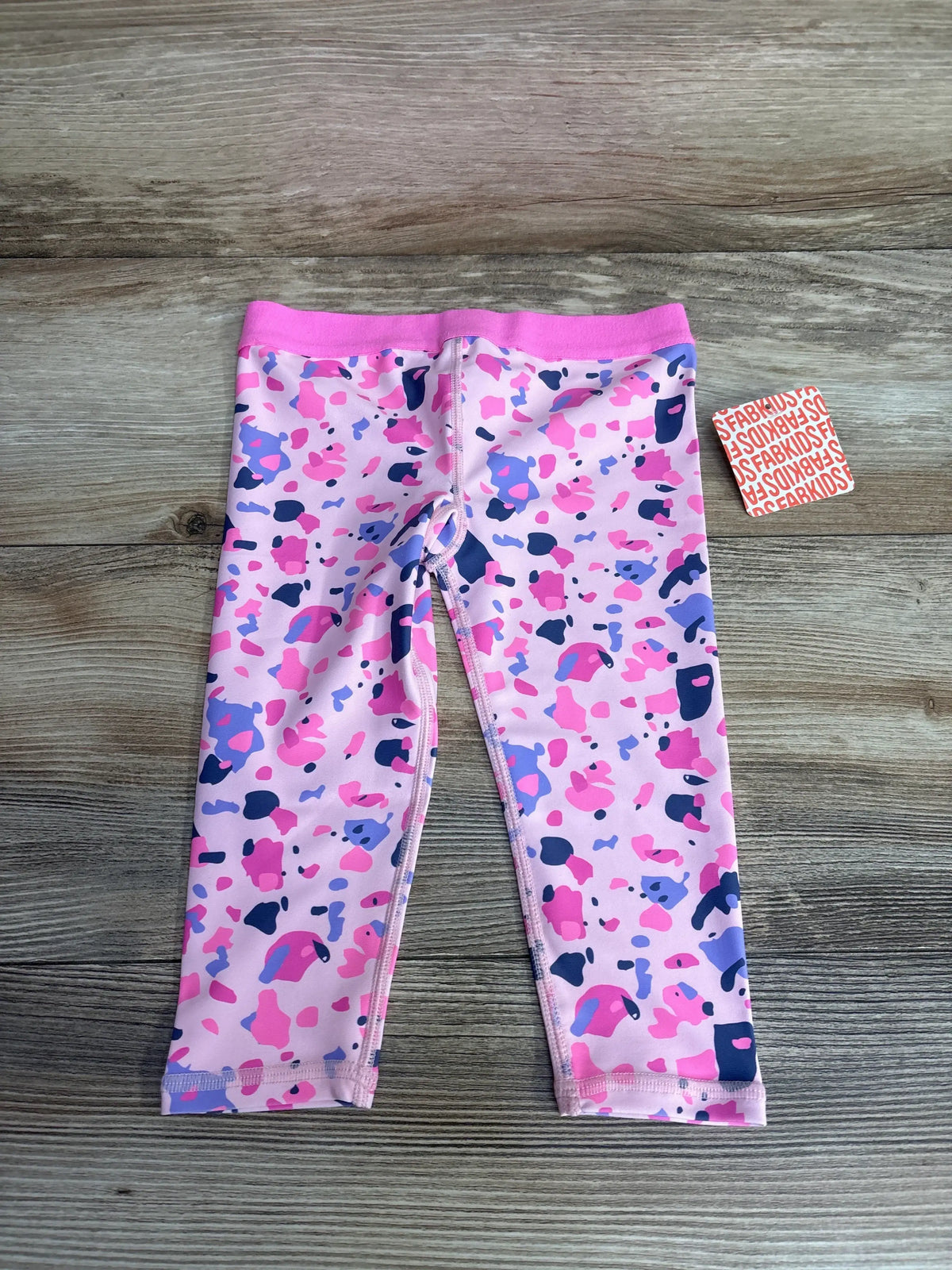 Front view of FabKids pink paint splatter leggings in size 2T with attached tag on a wood background