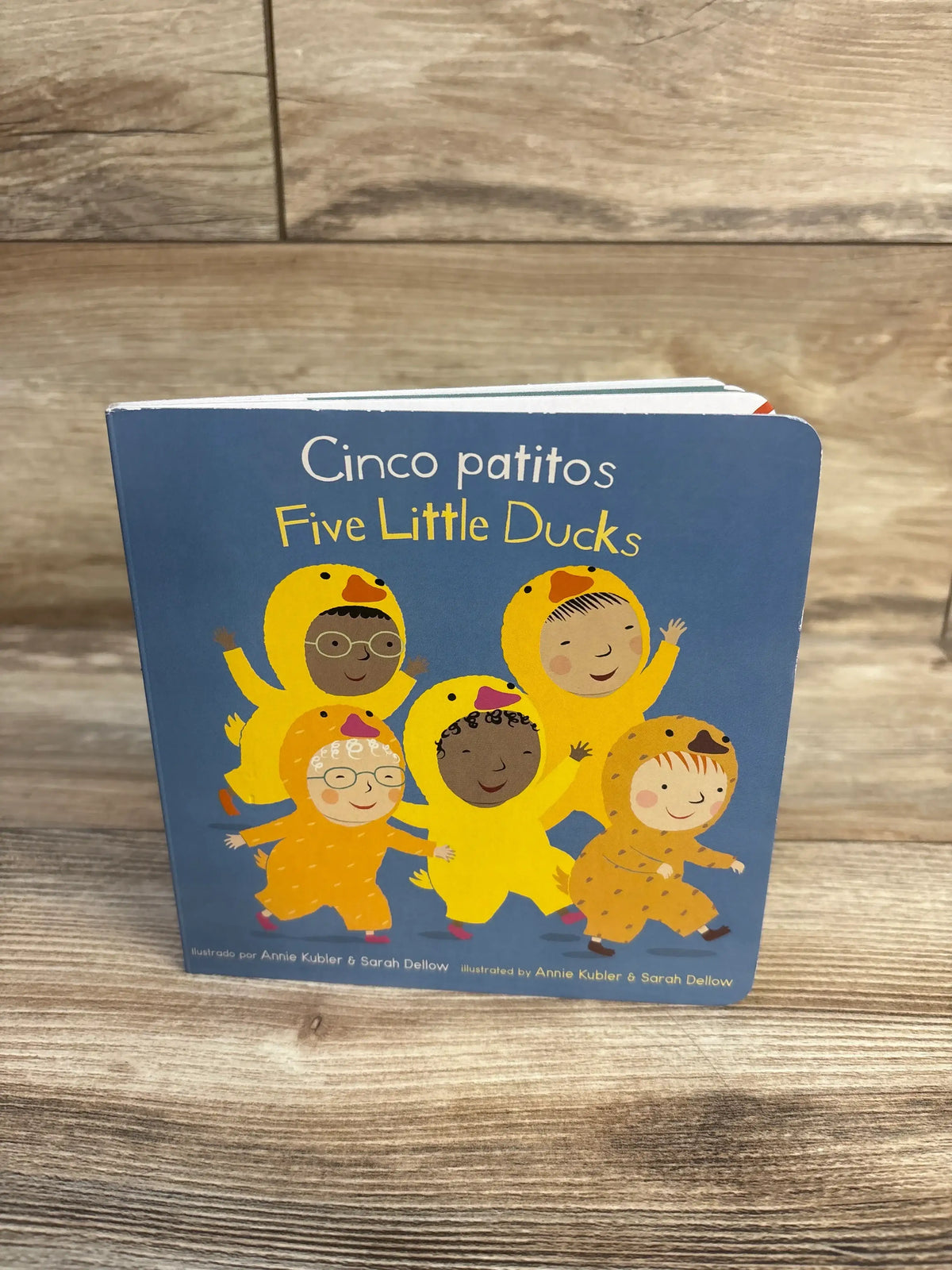 Front view of Cinco Patitos / Five Little Ducks board book showcasing five duck-costumed children on a wood background