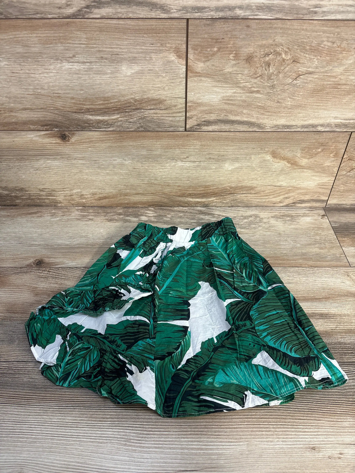 Overhead view of green tropical skirt, size 2T, showcasing vibrant leaf print and elastic waistband on a wood-look floor