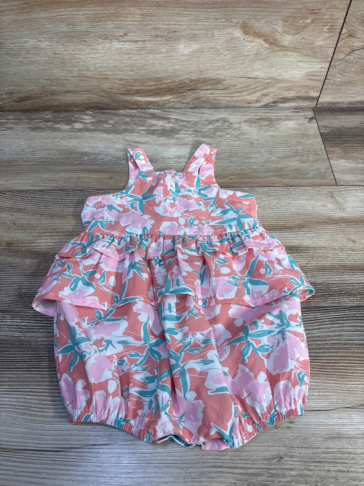 Front flat-lay of baby shortie romper showcasing ruffled peplum, floral print and elastic leg openings on wood background