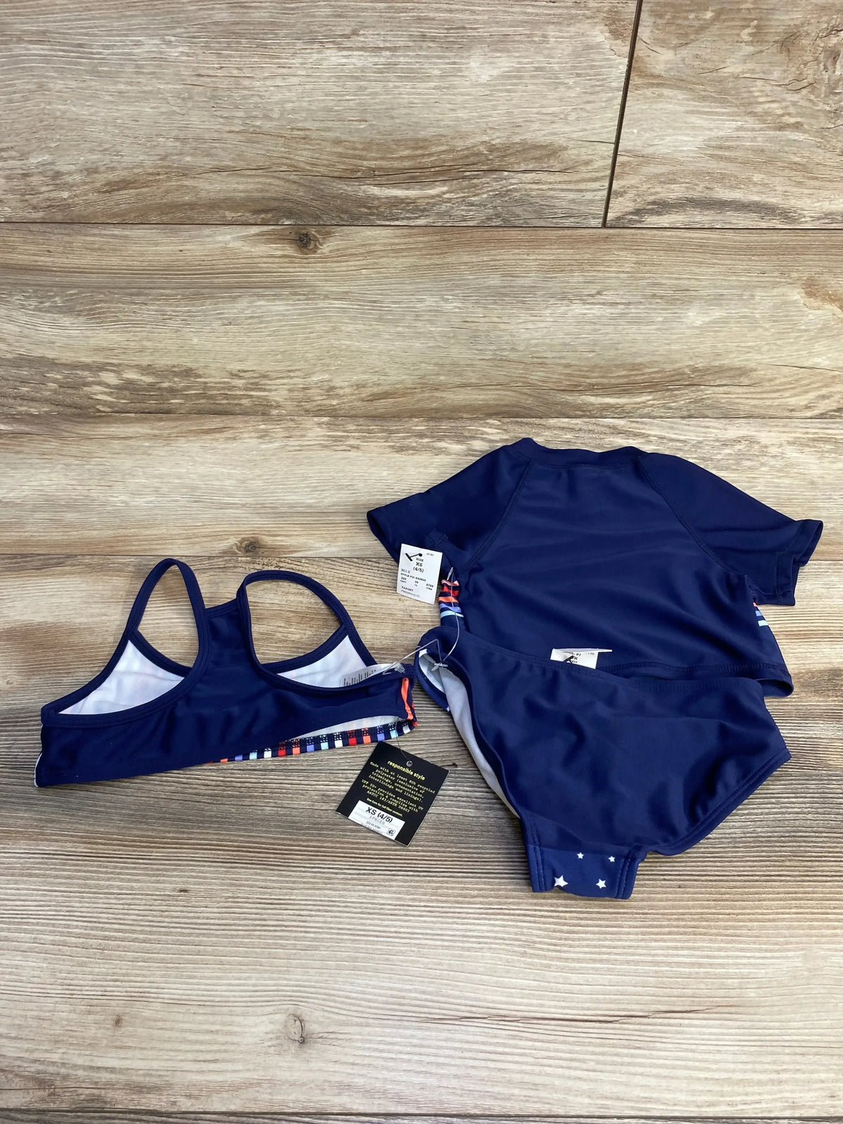 Flat lay of navy 3-piece toddler rashguard swim set: short-sleeve top, bralette top, and matching bottoms on wood floor