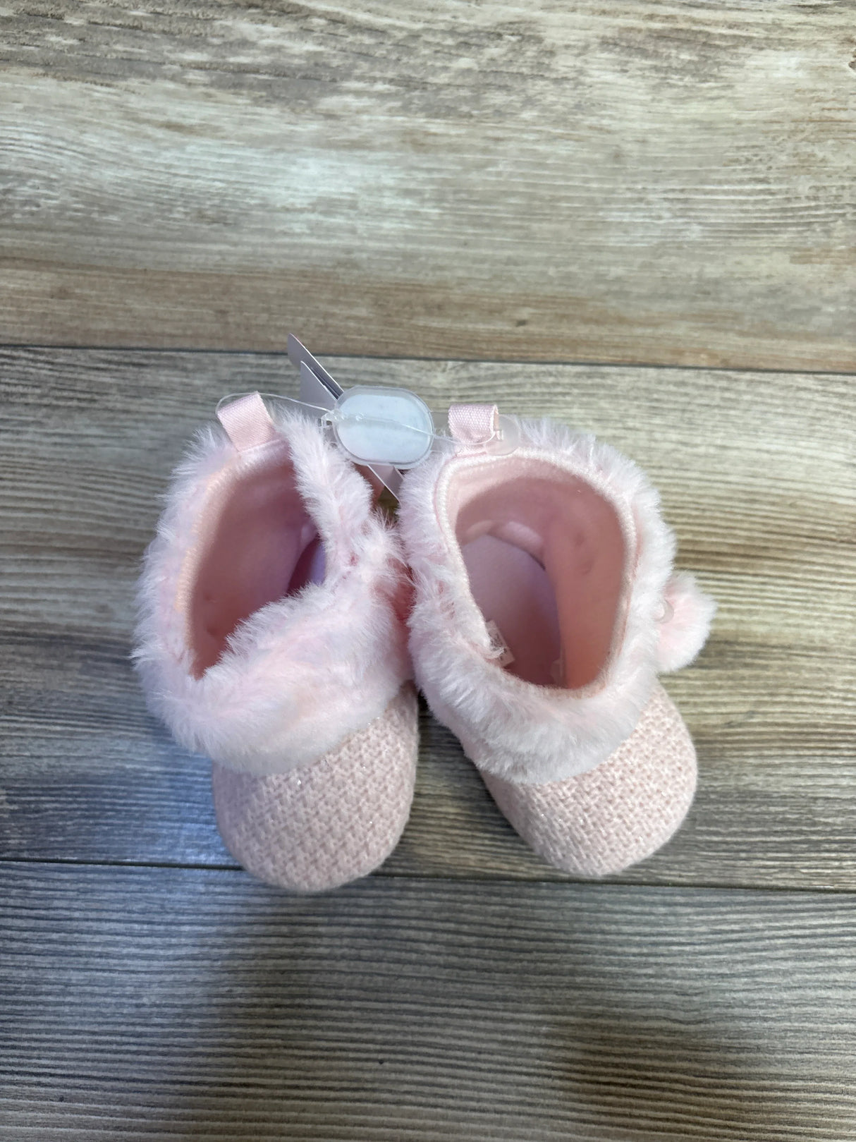 Top-down view of pink knit baby booties with faux-fur cuff and pom-pom detail on wood plank floor