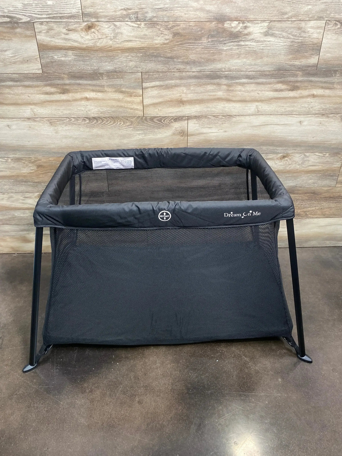 Front view of NEW Dream On Me Travel Light Playard in black showcasing mesh sides and padded rim on concrete floor