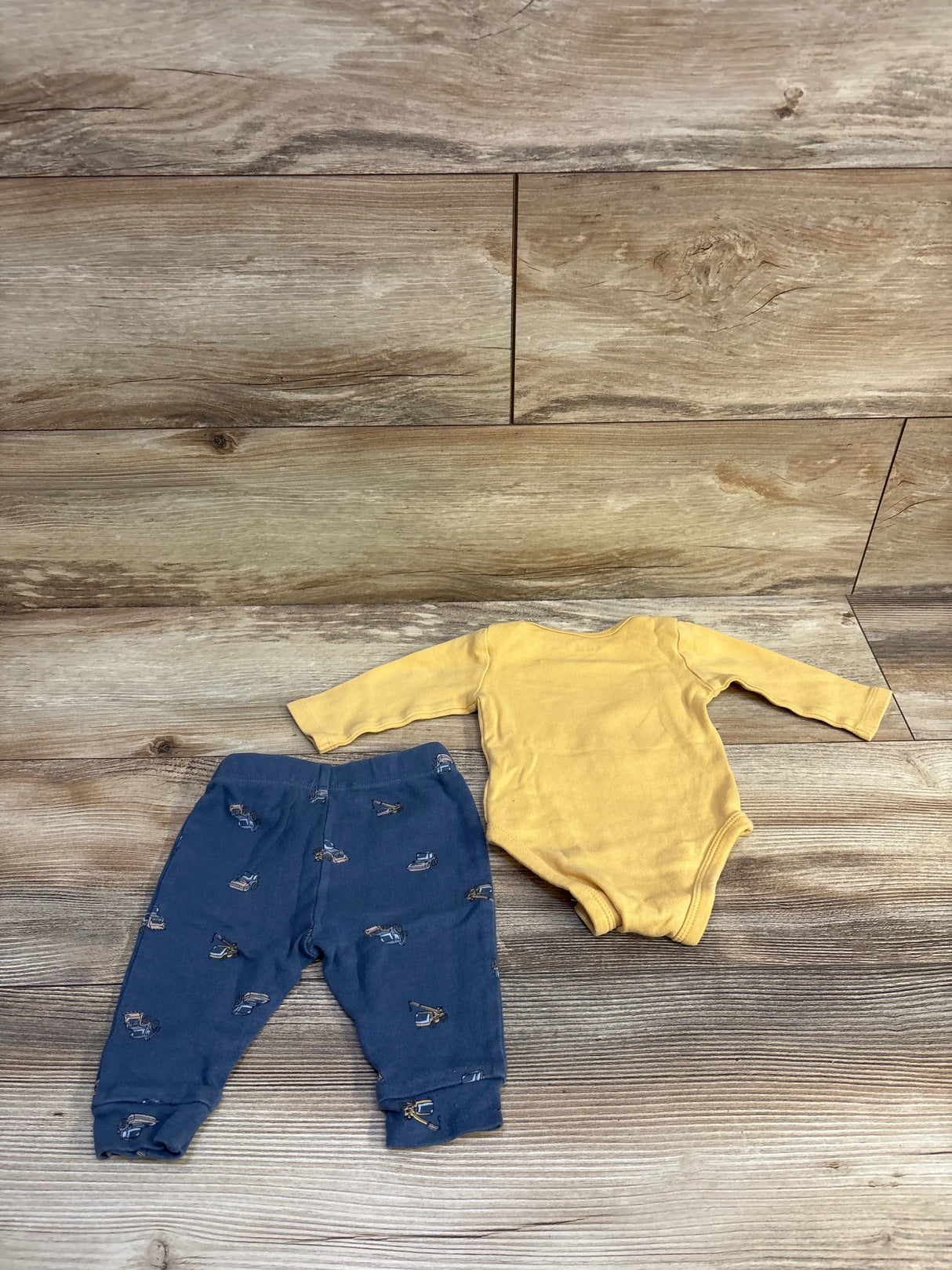 Flat lay of Pekkle 2-piece yellow long-sleeve bodysuit and navy bottoms with construction vehicle print on wood plank background