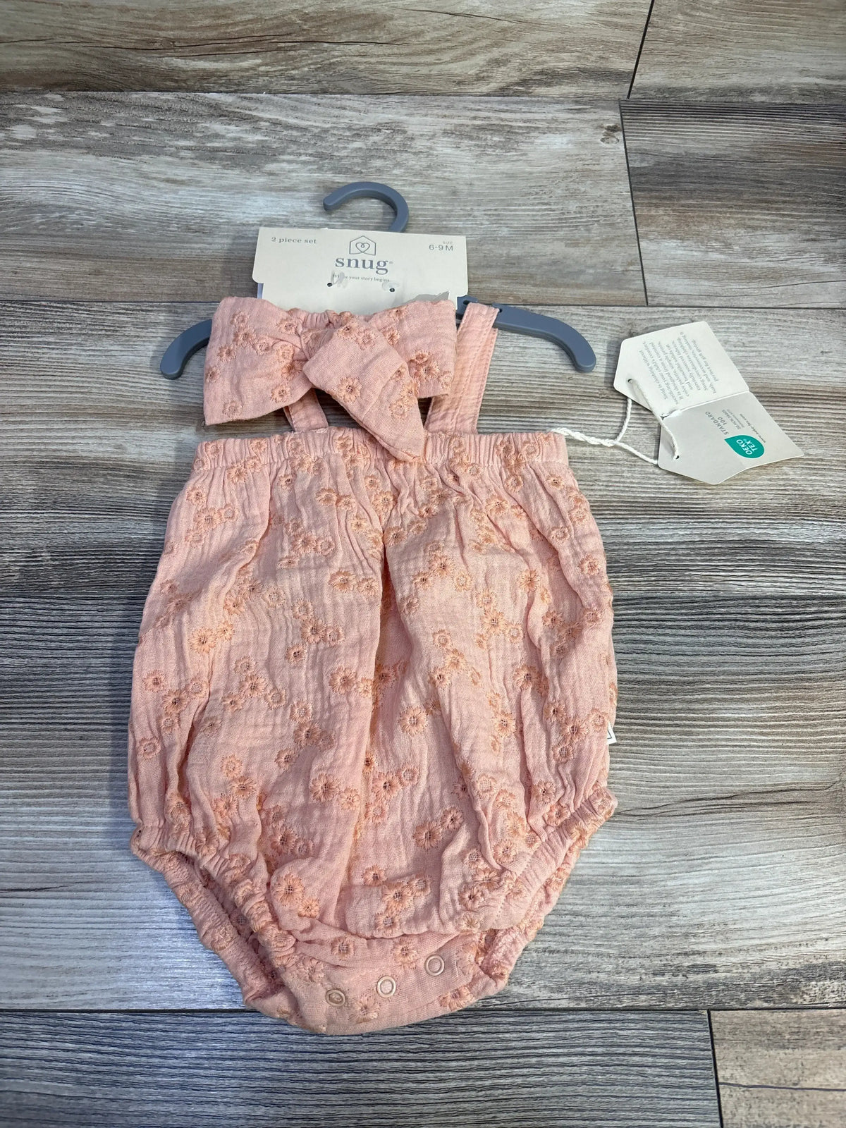 Front view of peach bubble romper with matching headband, floral embroidery, smocked straps and snap crotch on wood background