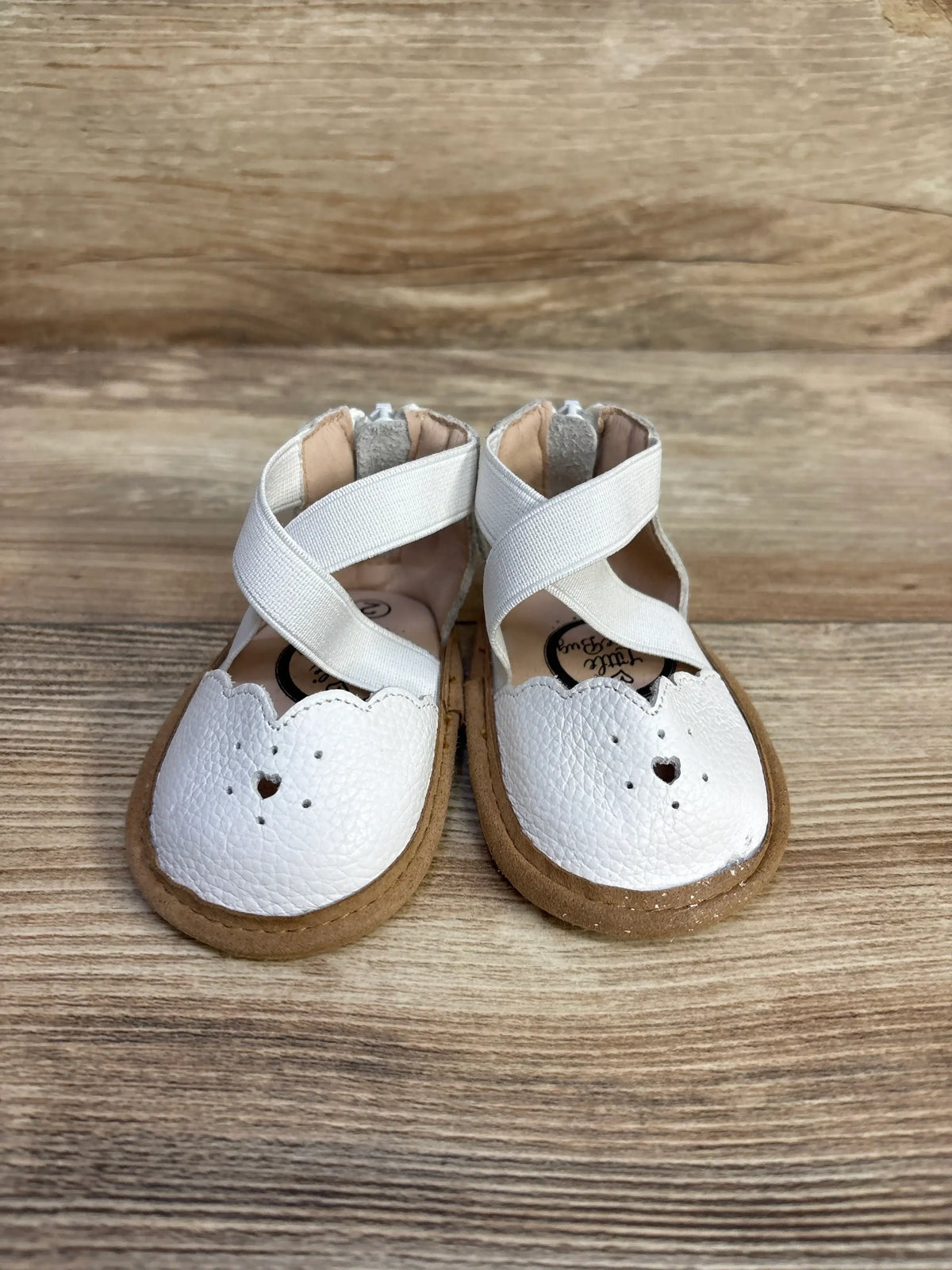 Front view of Sophia baby shoes in white with scalloped toe and heart cutout, crisscross elastic straps on wood background