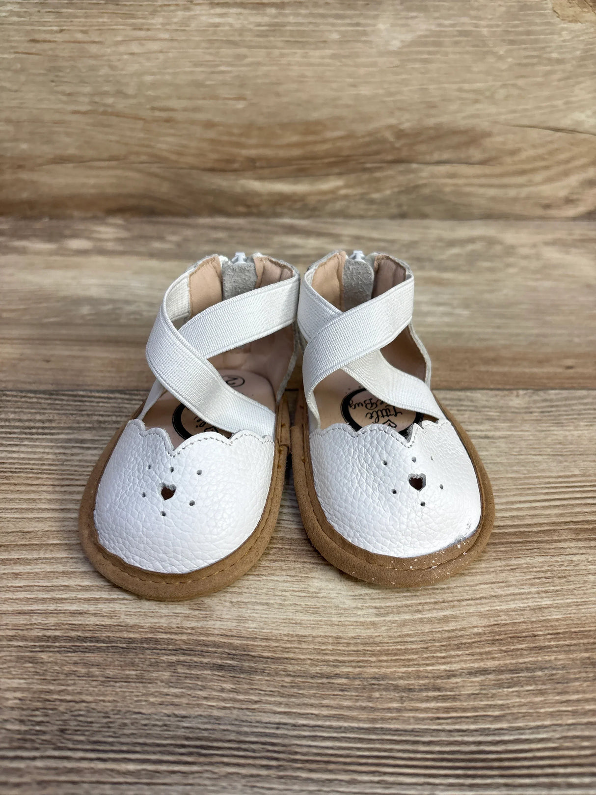 Front view of Sophia baby shoes in white with scalloped toe and heart cutout, crisscross elastic straps on wood background