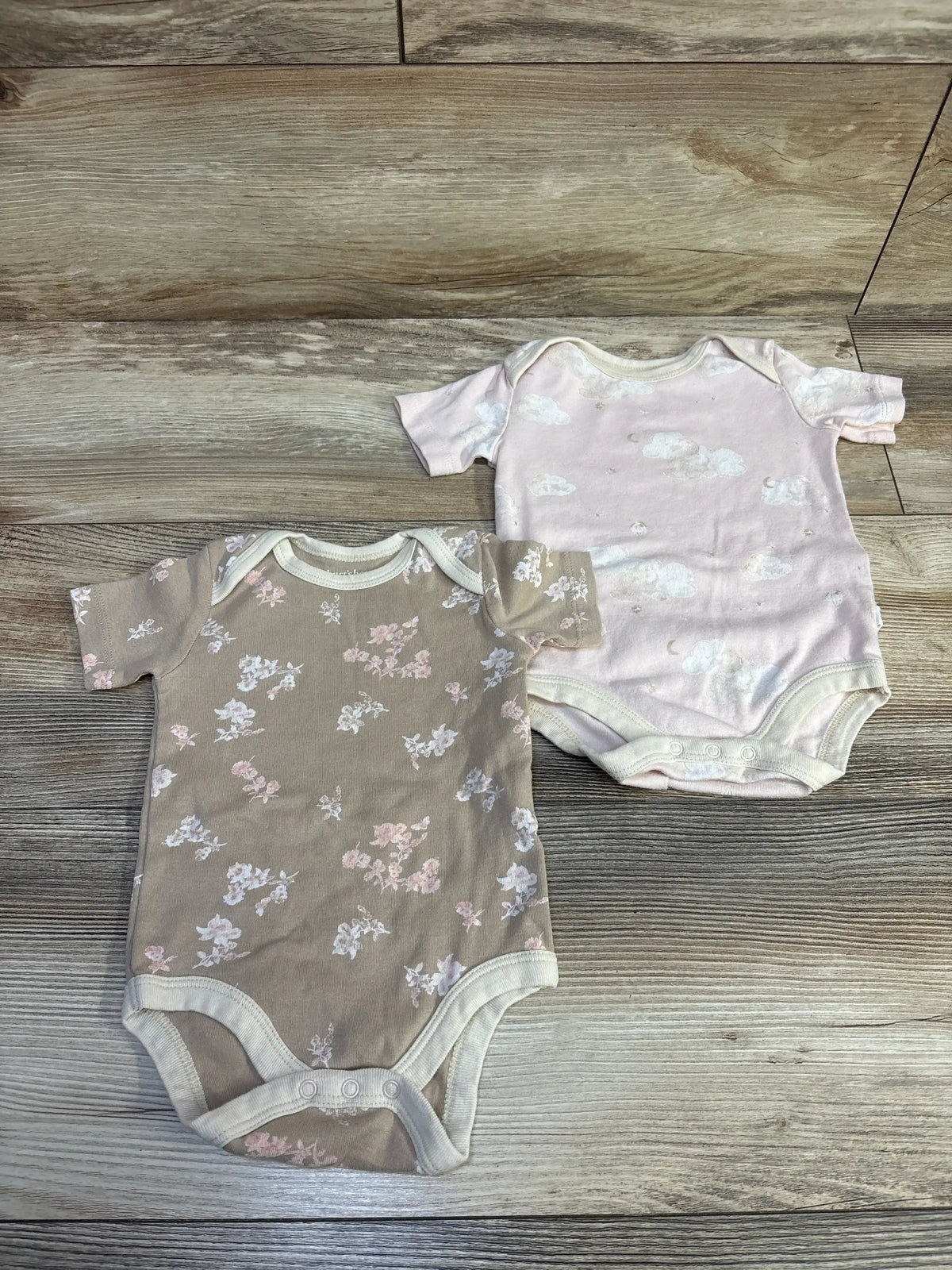 Top-down view of Burts Bees Baby 2pk bodysuits showcasing pink cloud and taupe floral prints, short sleeves, and snap closures